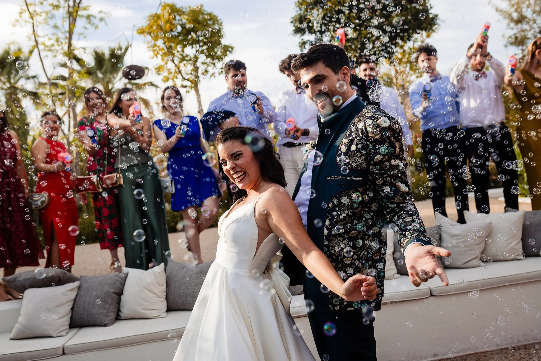 Novios bailando en una boda al aire libre, rodeados de amigos y familiares lanzando burbujas, en un ambiente alegre y festivo.