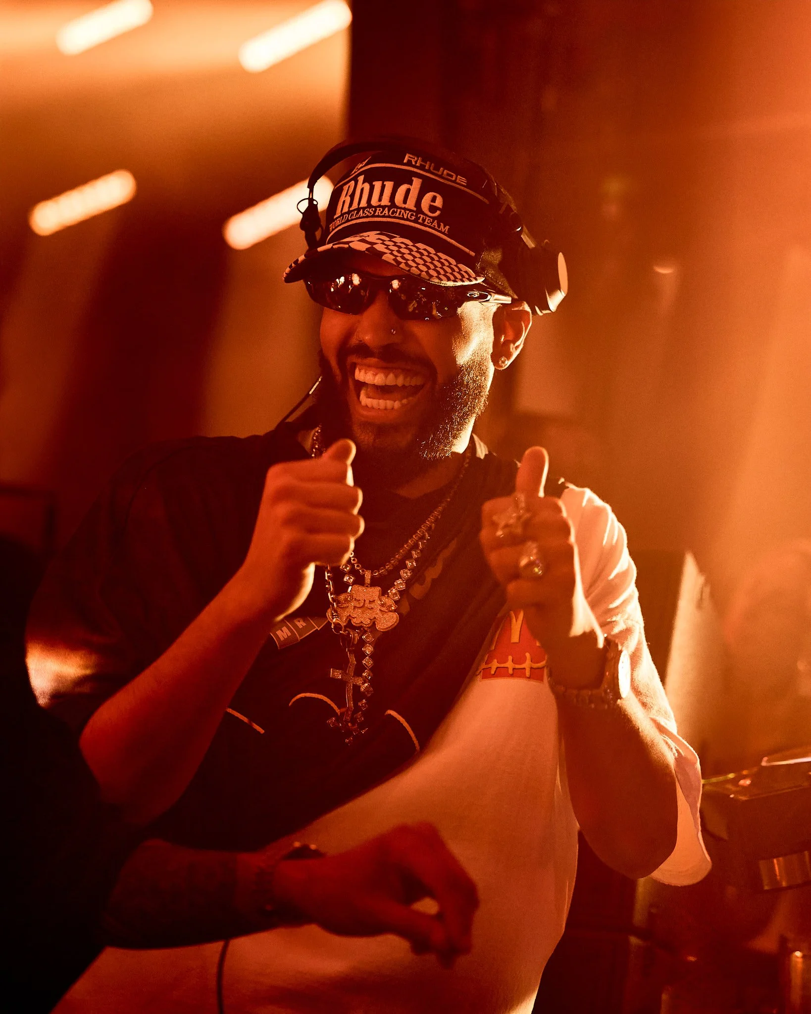 Hombre sonriendo con gafas de sol, gorra y varios collares, mostrando una señal de pulgar arriba en un ambiente con luz anaranjada.
