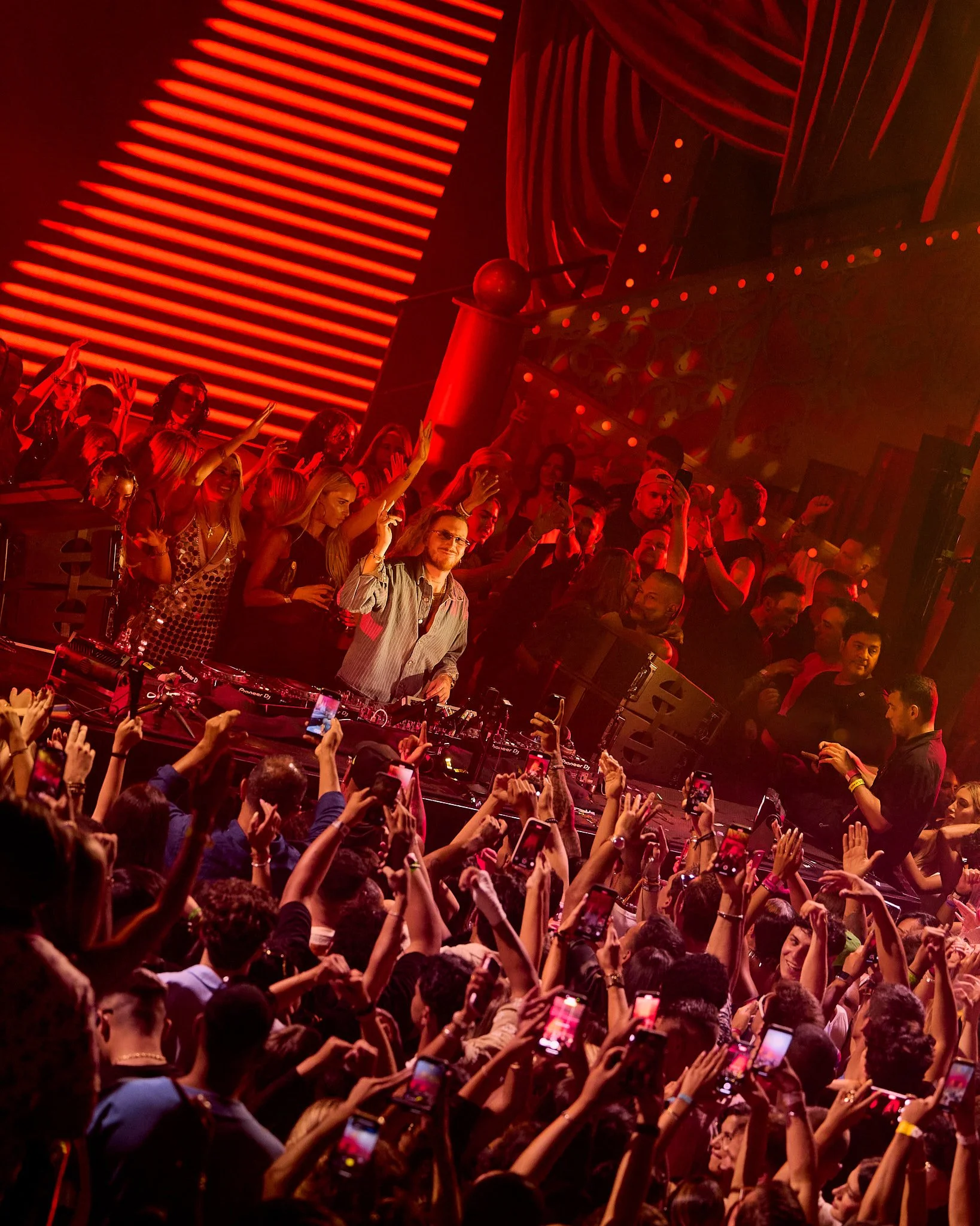 Una multitud en un concierto o fiesta con una pista de DJ en el centro y un escenario iluminado en tonos rojos, con personas bailando y tomando fotos con sus teléfonos móviles.