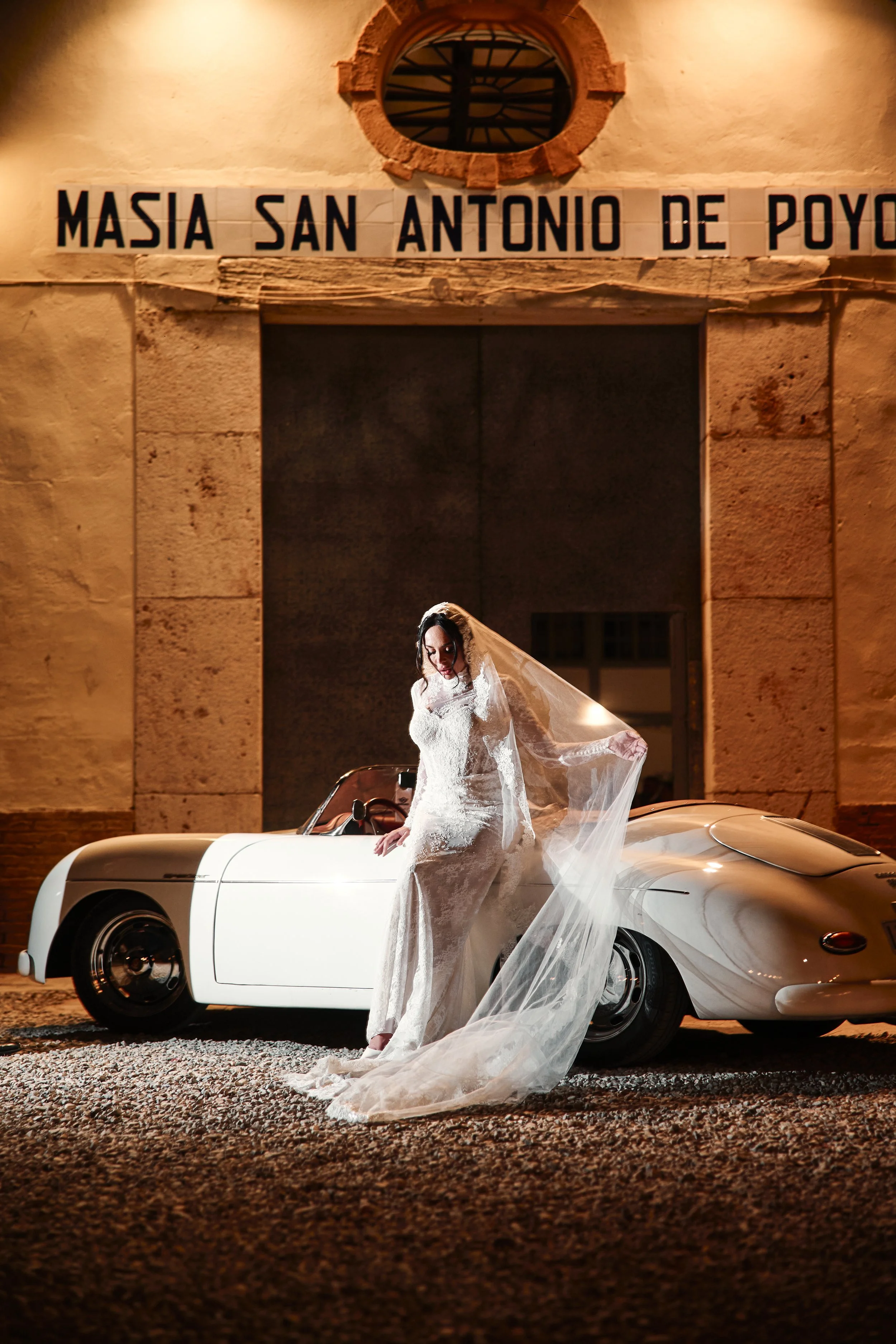 Mujer con vestido de novia posando en un coche clásico blanco, en un entorno urbano de noche, detrás de un edificio con cartel que dice "Masia San Antonio de Poyo".