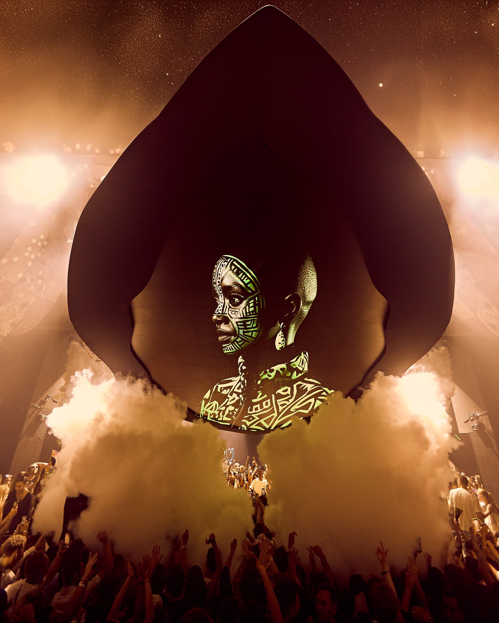 Una presentación en un escenario con una gran proyección de una mujer con maquillaje y accesorios tradicionales africanos, rodeada de humo y un público con las manos levantadas.