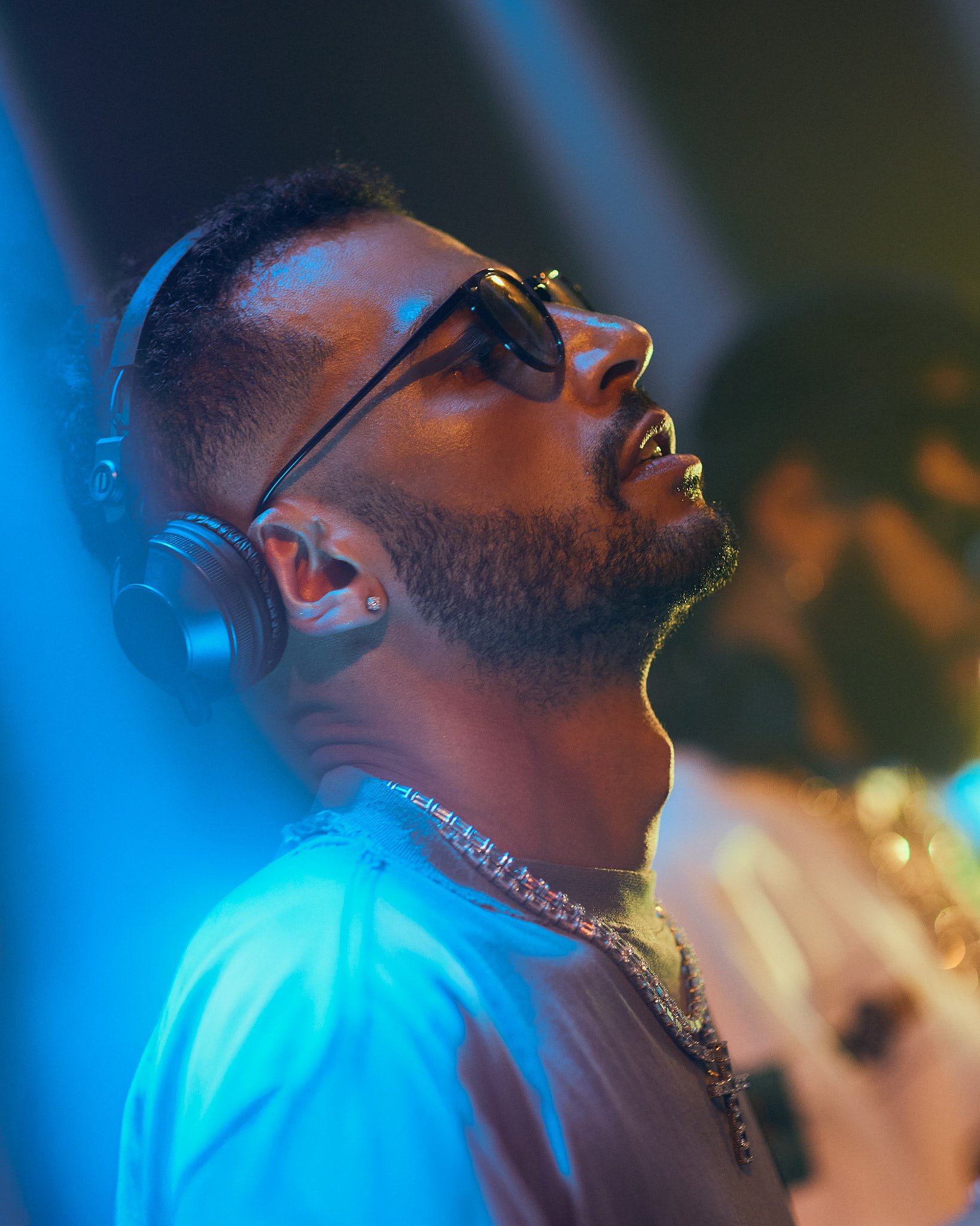 Joven con gafas oscuras, auriculares, y collar con cruz, disfrutando música en un ambiente con luces de neón, fondo desenfocado.