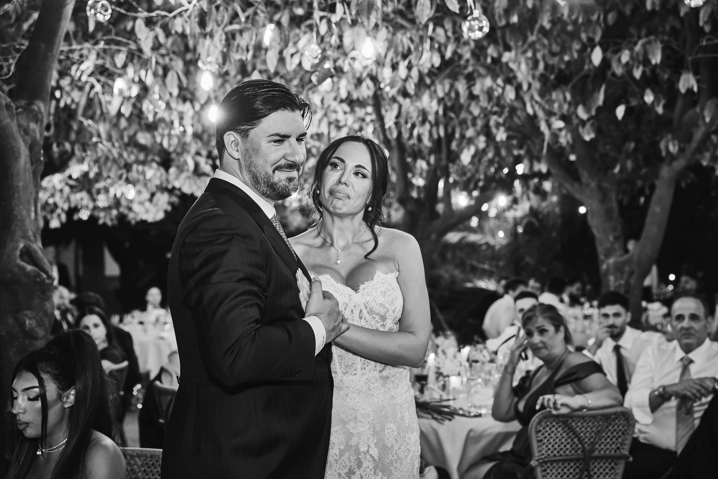 Pareja de novios en su boda, el hombre viste un traje oscuro y la mujer un vestido blanco de encaje, en una ceremonia al aire libre con árboles y luces decorativas en el fondo, acompañados por invitados.