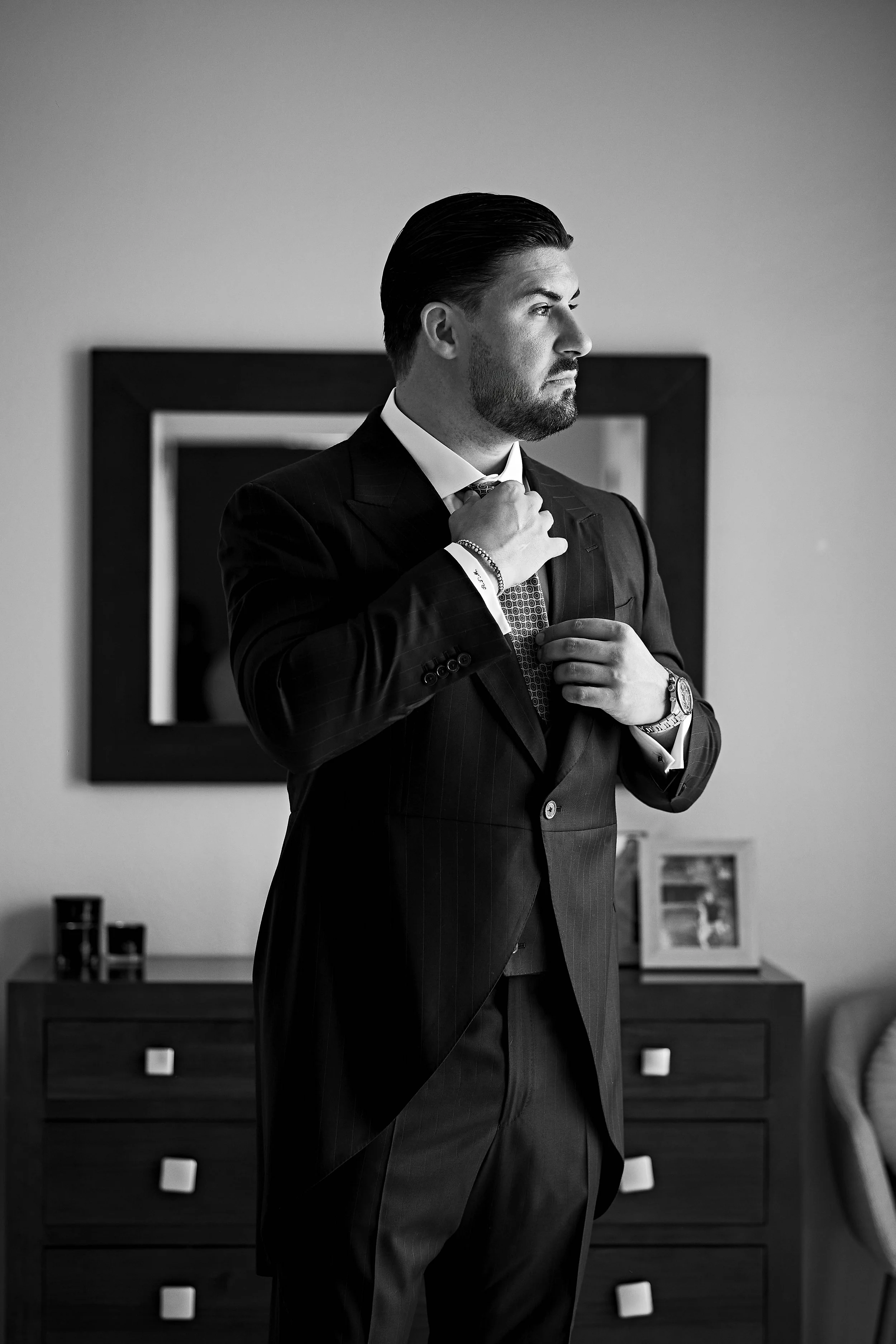 Hombre con barba y bigote, vestido con traje formal, ajustándose la corbata en un interior decorado con muebles y cuadros.