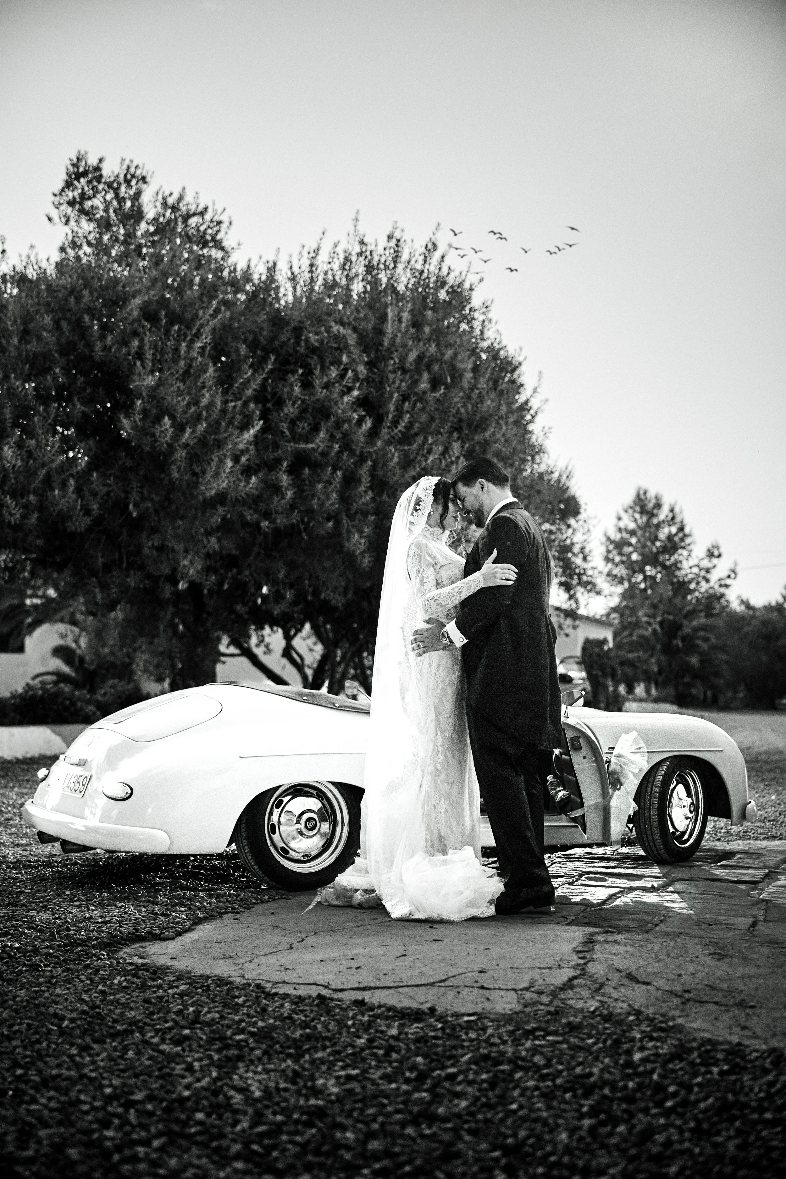 Pareja de novios besándose frente a un coche clásico, con árboles y aves en el cielo de fondo, en blanco y negro.