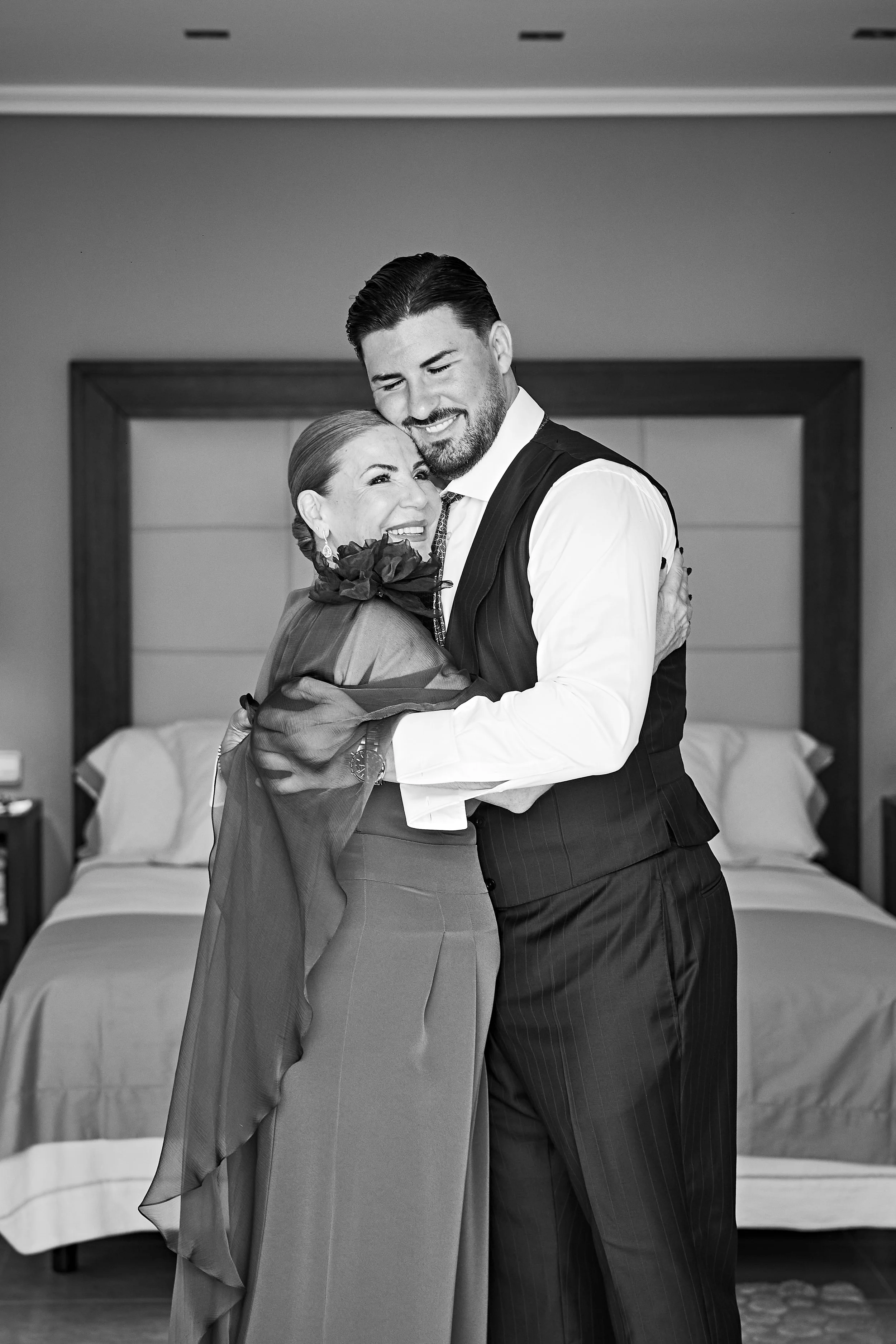 Una mujer mayor y un hombre joven se abrazan en una habitación, ambos sonriendo. La mujer lleva un vestido largo y un florero en el cabello, el hombre viste saco y camisa. Fondo con una cama y un cuadro en la pared.
