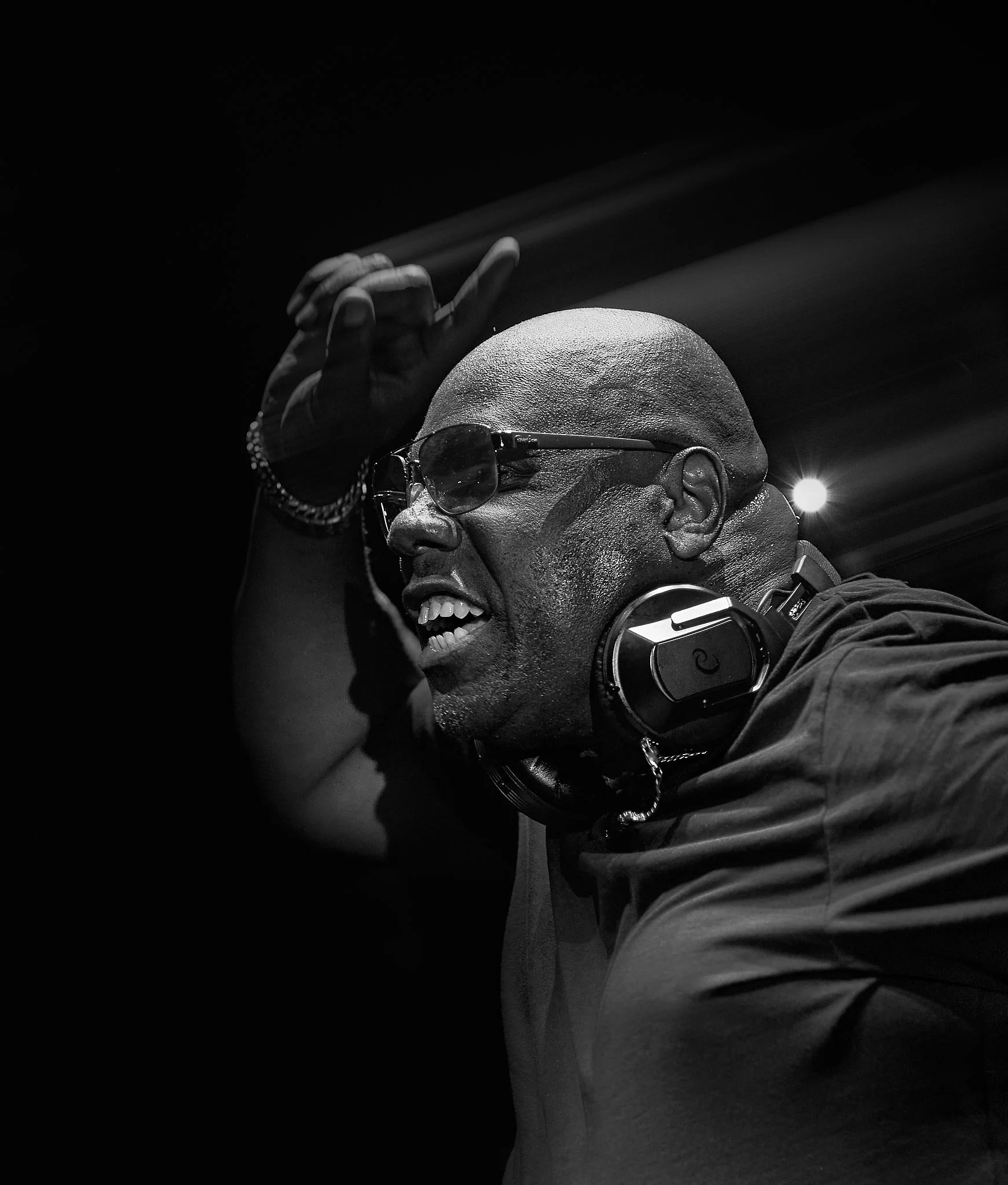 Carl Cox con gafas y auriculares, sonriendo y con la mano levantada, en un entorno oscuro, expresando alegría o entusiasmo.