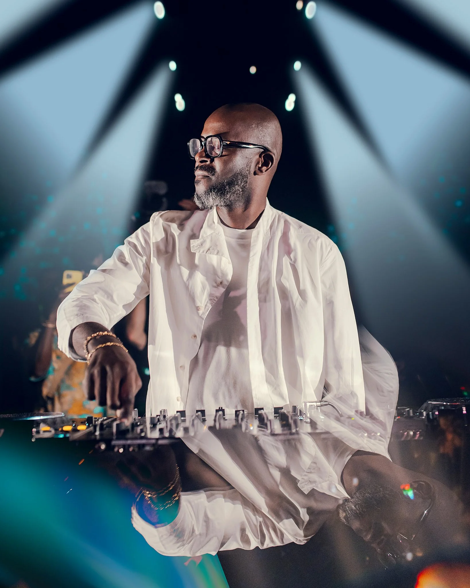 Hombre con barba y gafas de sol, vestido con ropa blanca, mezclando música en un controlador de DJ en un escenario con luces y ambiente de fiesta.