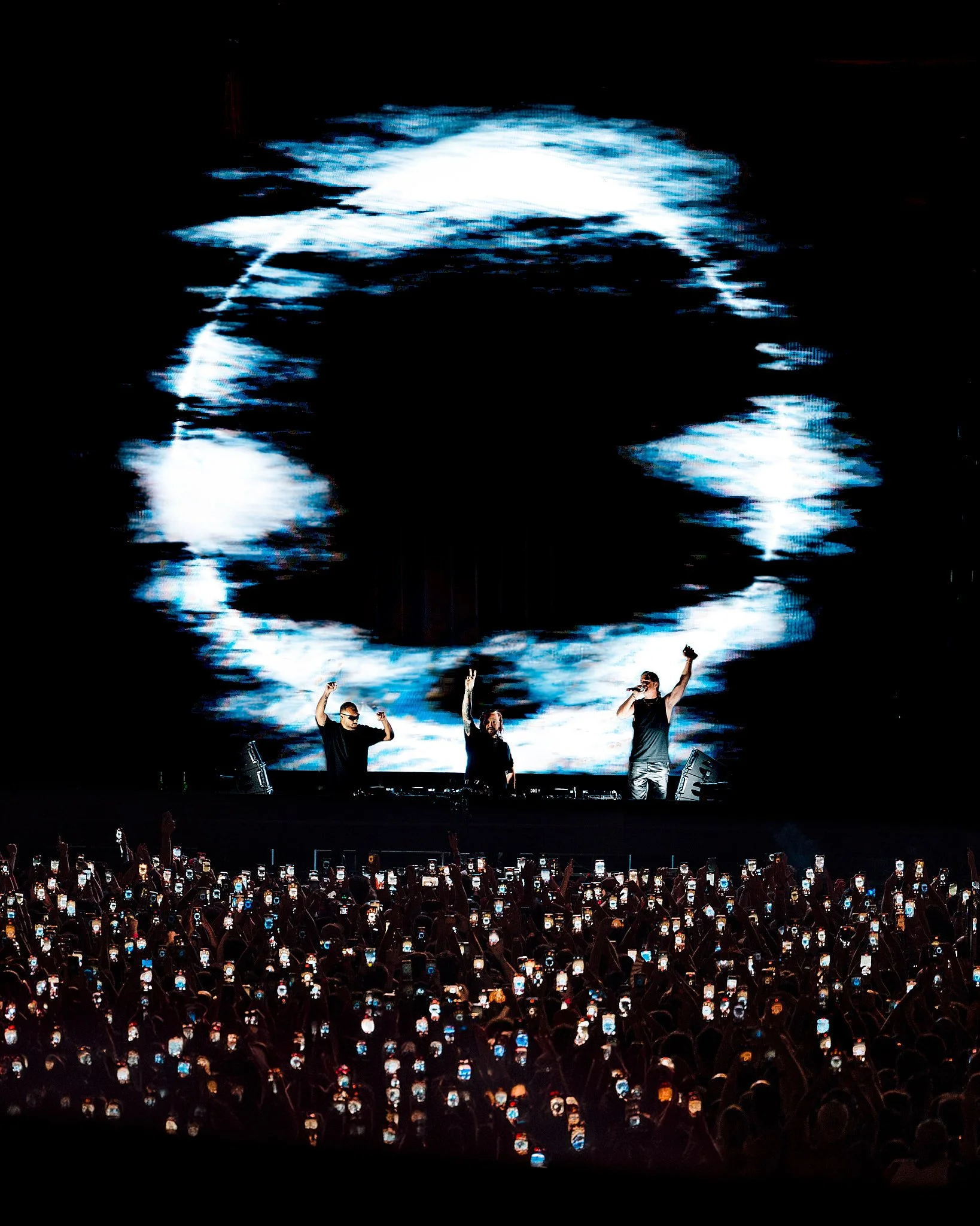 Concierto en vivo con tres artistas en el escenario, frente a una multitud que sostiene teléfonos, y una gran pantalla que muestra una imagen estilizada del rostro de un hombre.