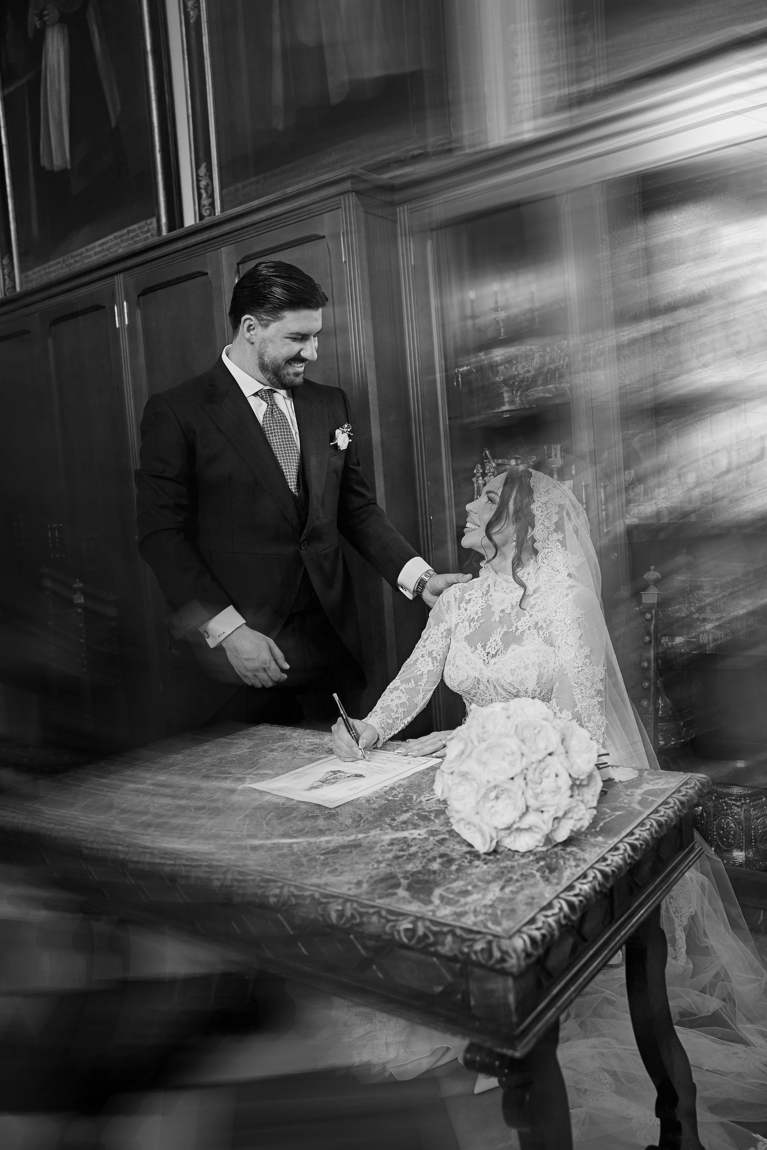 Una pareja en una boda firma un documento sobre una mesa antigua con un ramo de flores blancas. La escena es en blanco y negro, en un ambiente elegante con madera y decoraciones clásicas.