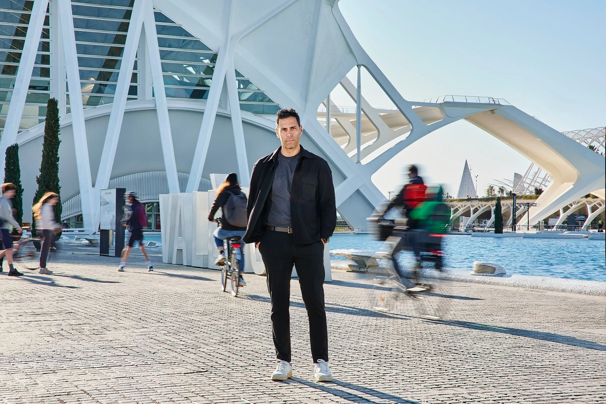 Hombre de pie frente a un edificio moderno y arquitectónico, con personas caminando y en bicicleta en el fondo cerca del agua.