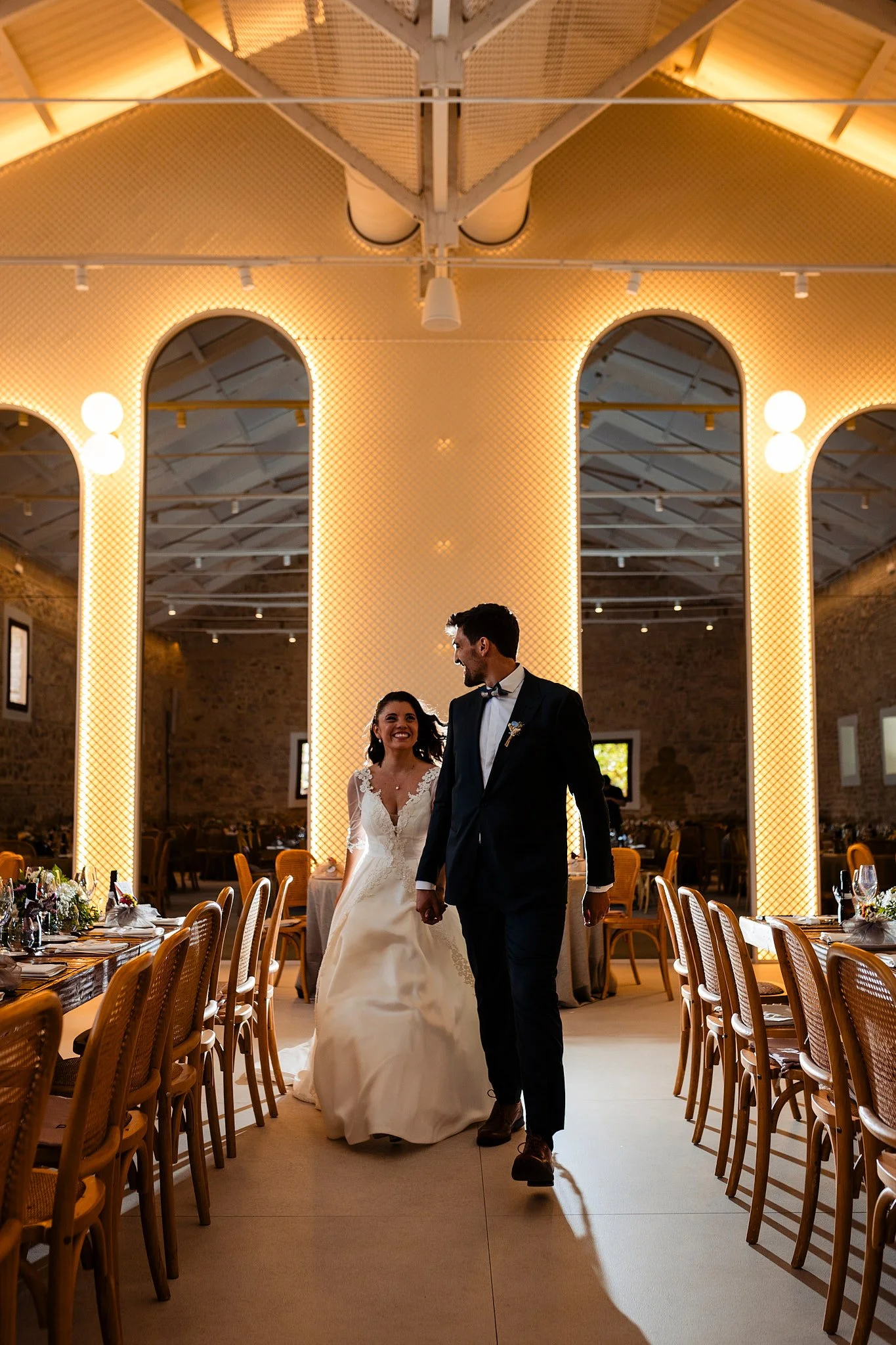 Pareja de novios en una boda, caminando en un salón elegantemente decorado con iluminación cálida y mesas arregladas para la celebración.