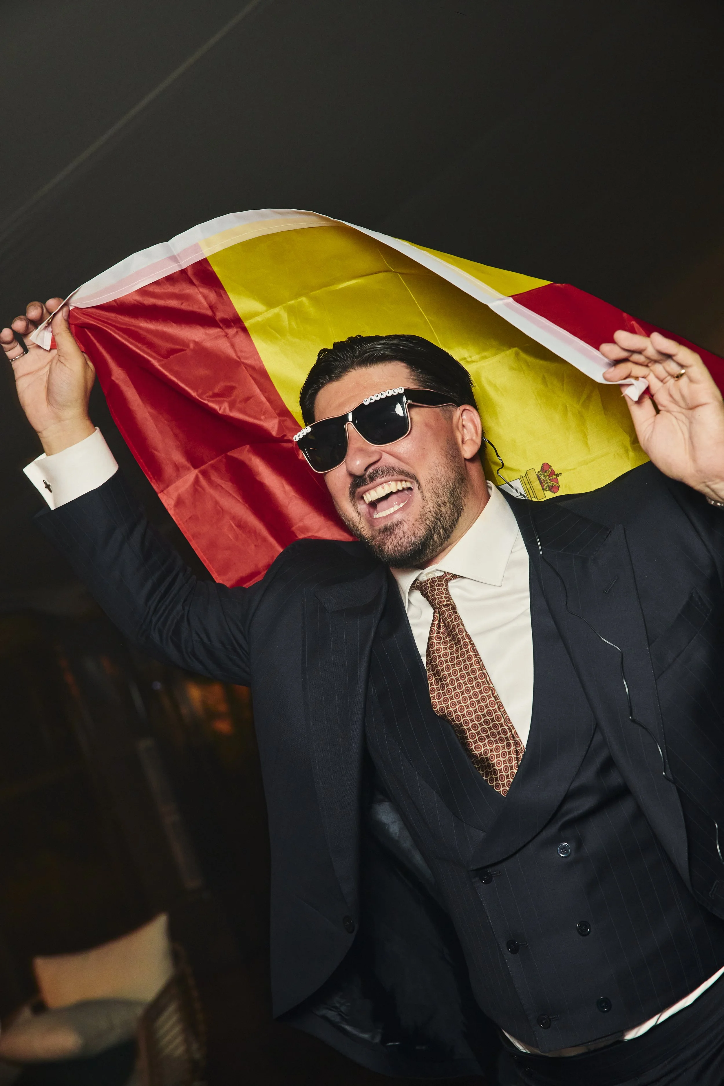 Hombre con gafas oscuras y barba, vestido con etiqueta de matrimonio, sostiene una bandera de España y sonríe en un evento.