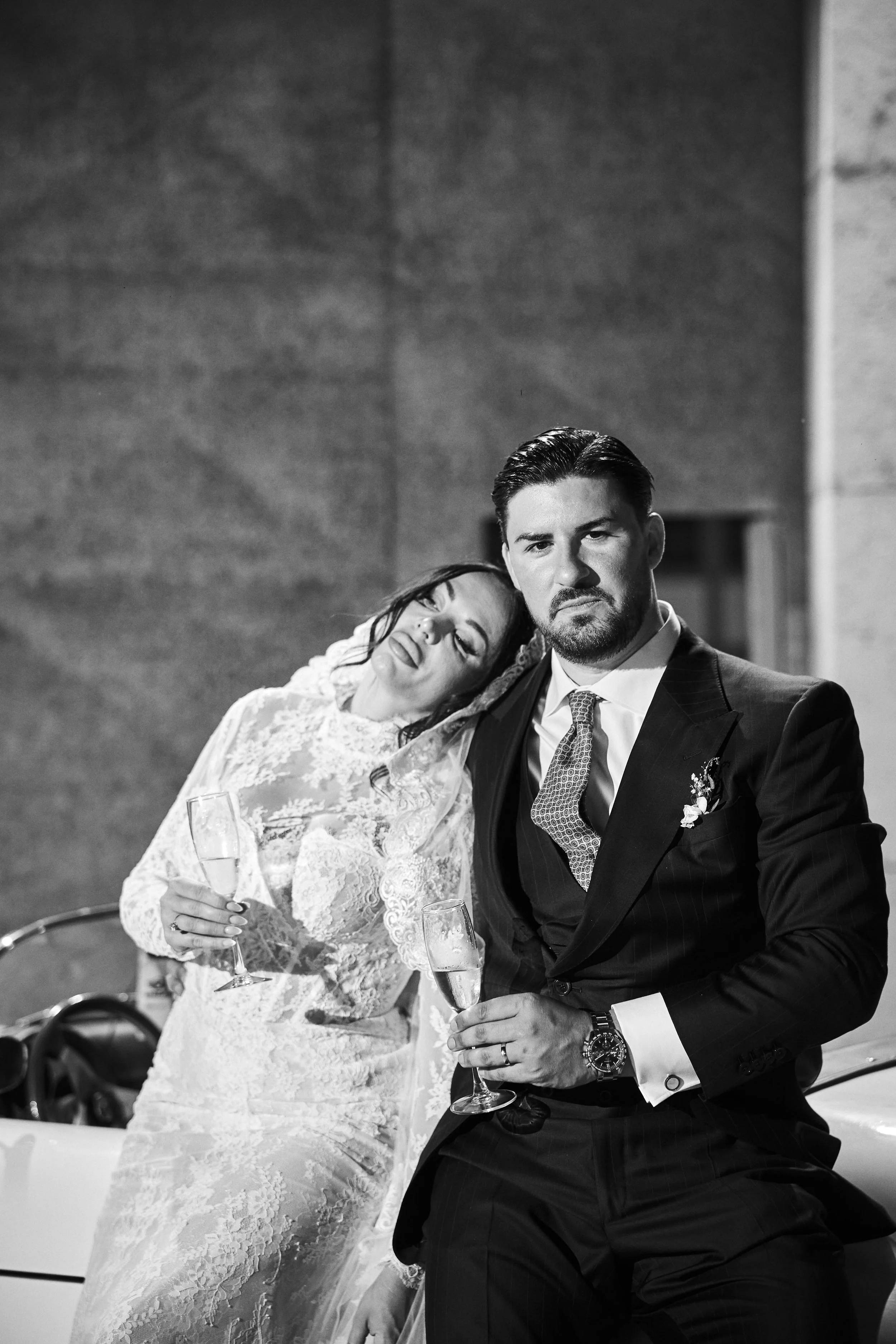 Pareja de novios en una boda, la mujer apoya su cabeza en el hombro del hombre mientras sostiene una copa de champagne, en un entorno elegante en blanco y negro.