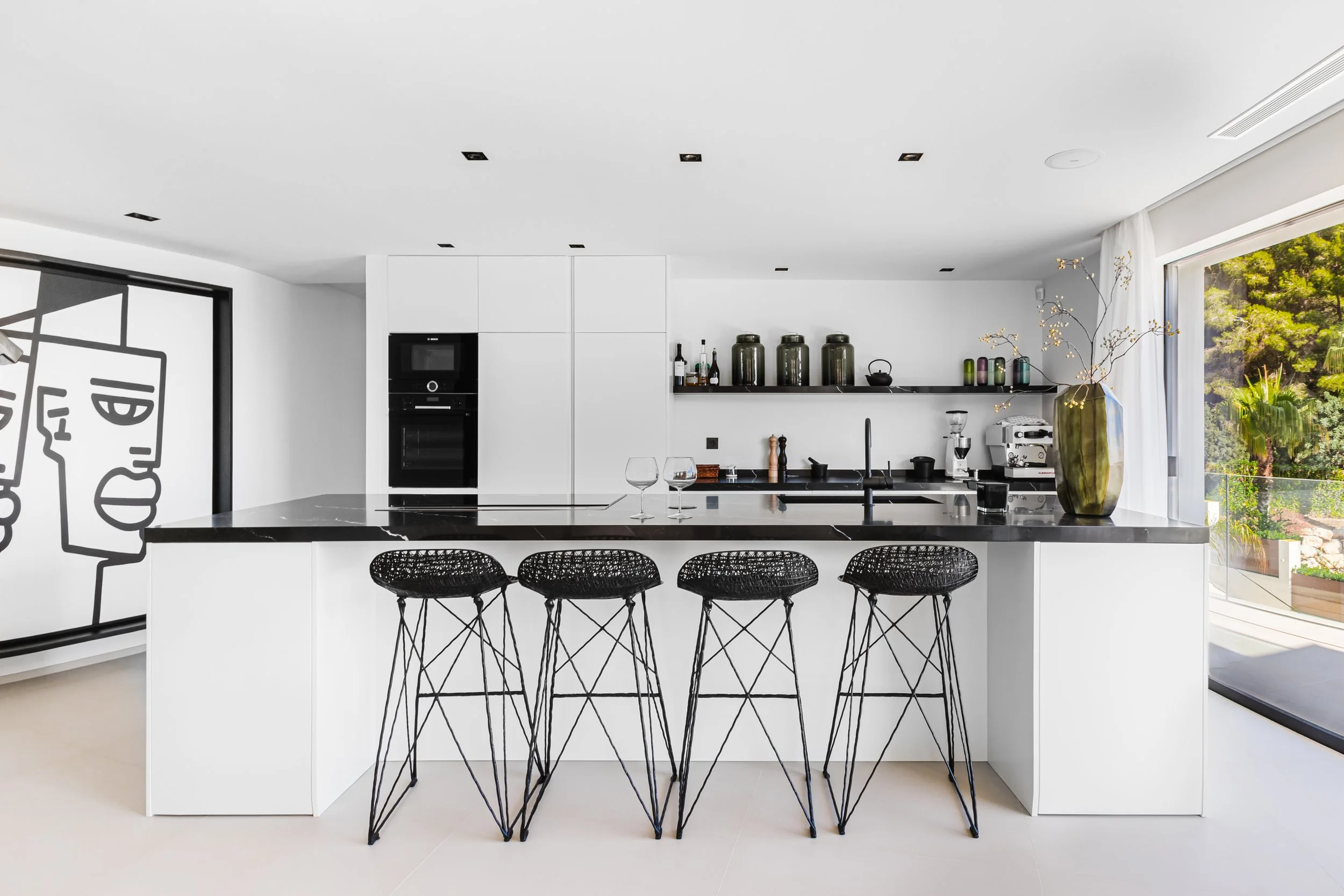 Cocina moderna con barra blanca, sillas negras de mimbre y vista a un jardín con vegetación