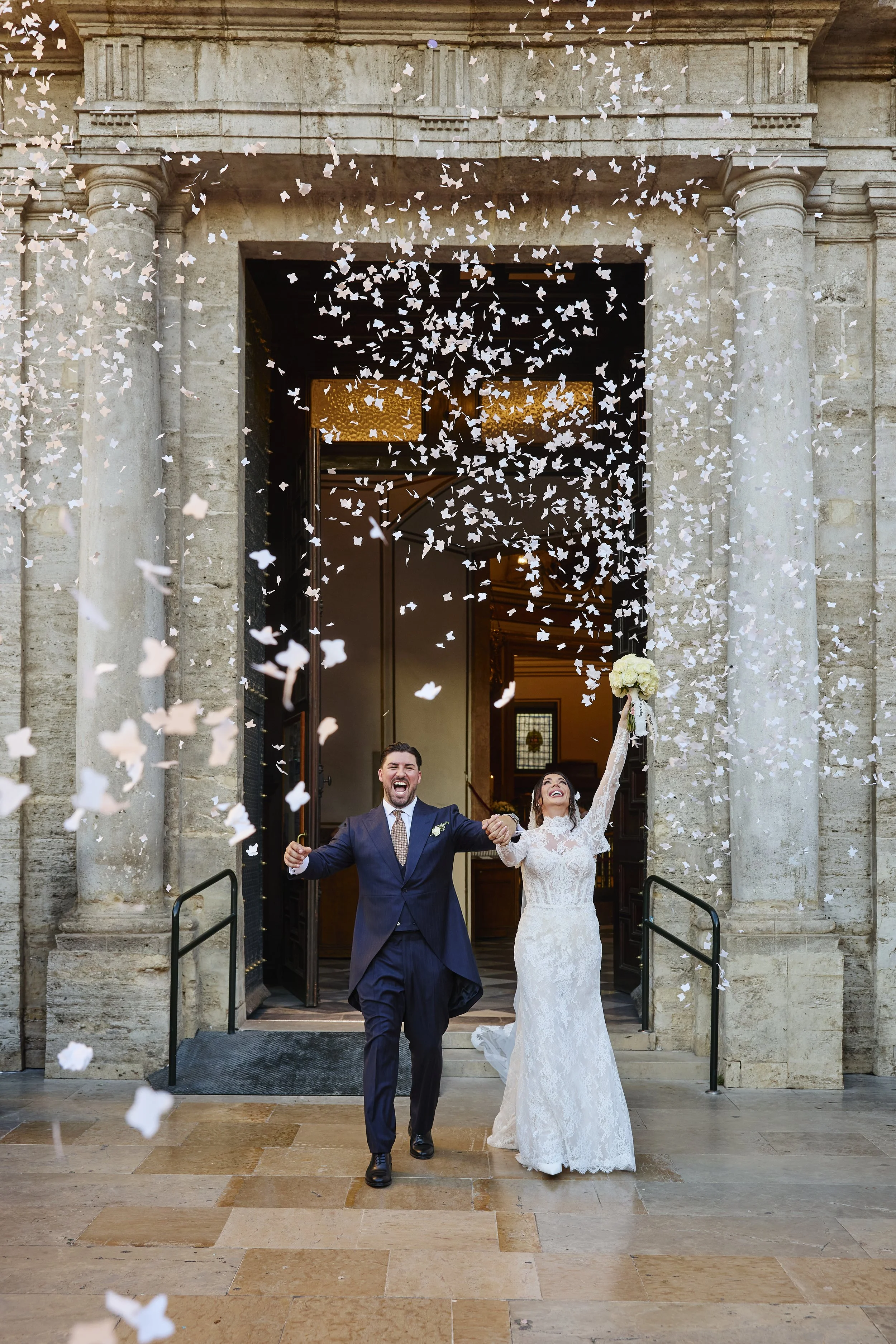 Una pareja de recién casados celebra en la entrada de una iglesia, con pétalos de flores cayendo, ella en vestido de novia y él en traje azul oscuro, ambos sonriendo y luciendo felices