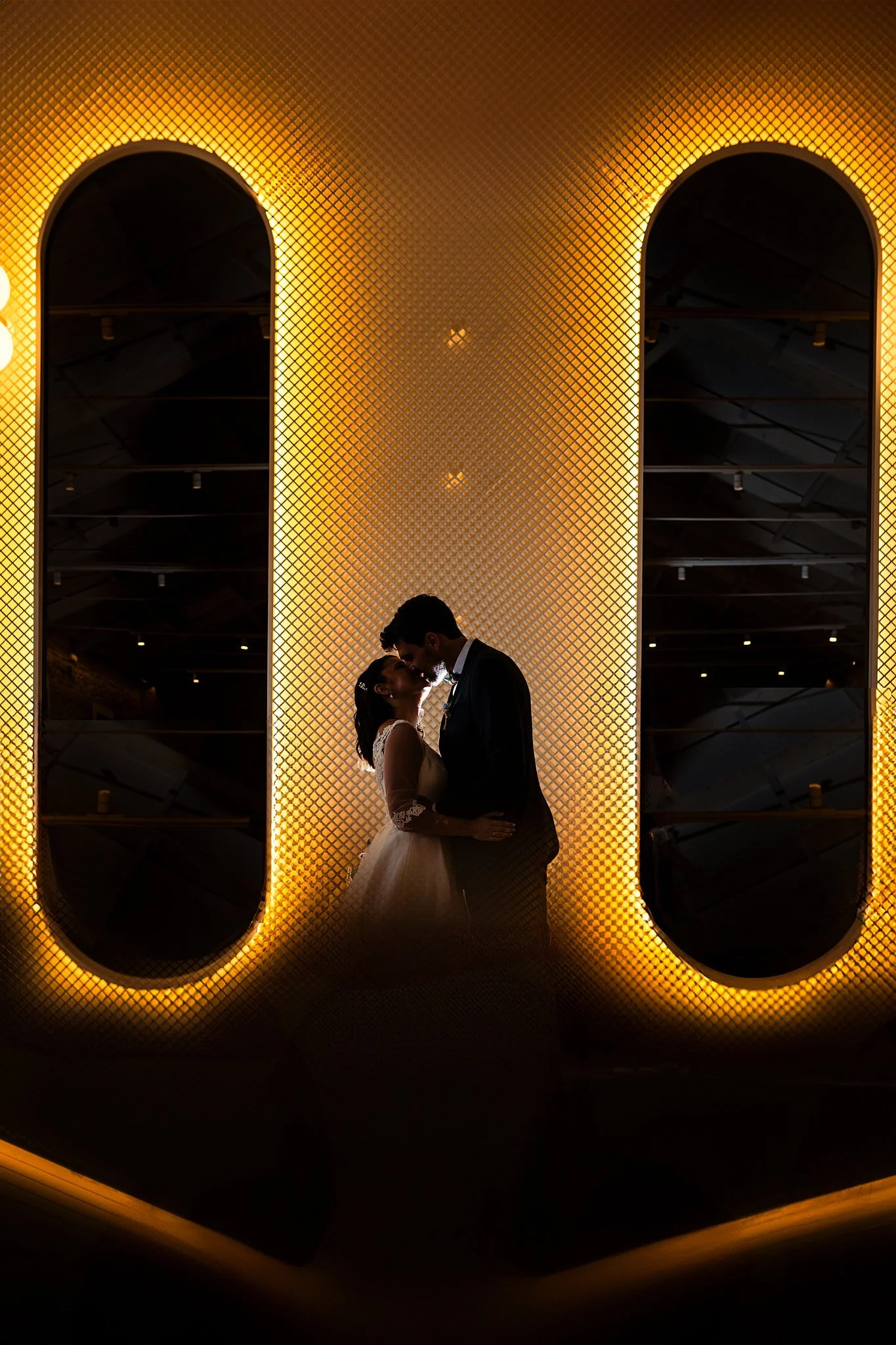 Pareja de novios en un momento romántico, de espaldas, con el hombre usando traje y la mujer vestido de novia, en un fondo iluminado con luces doradas y una pared decorativa con ventanas ovaladas oscuras.