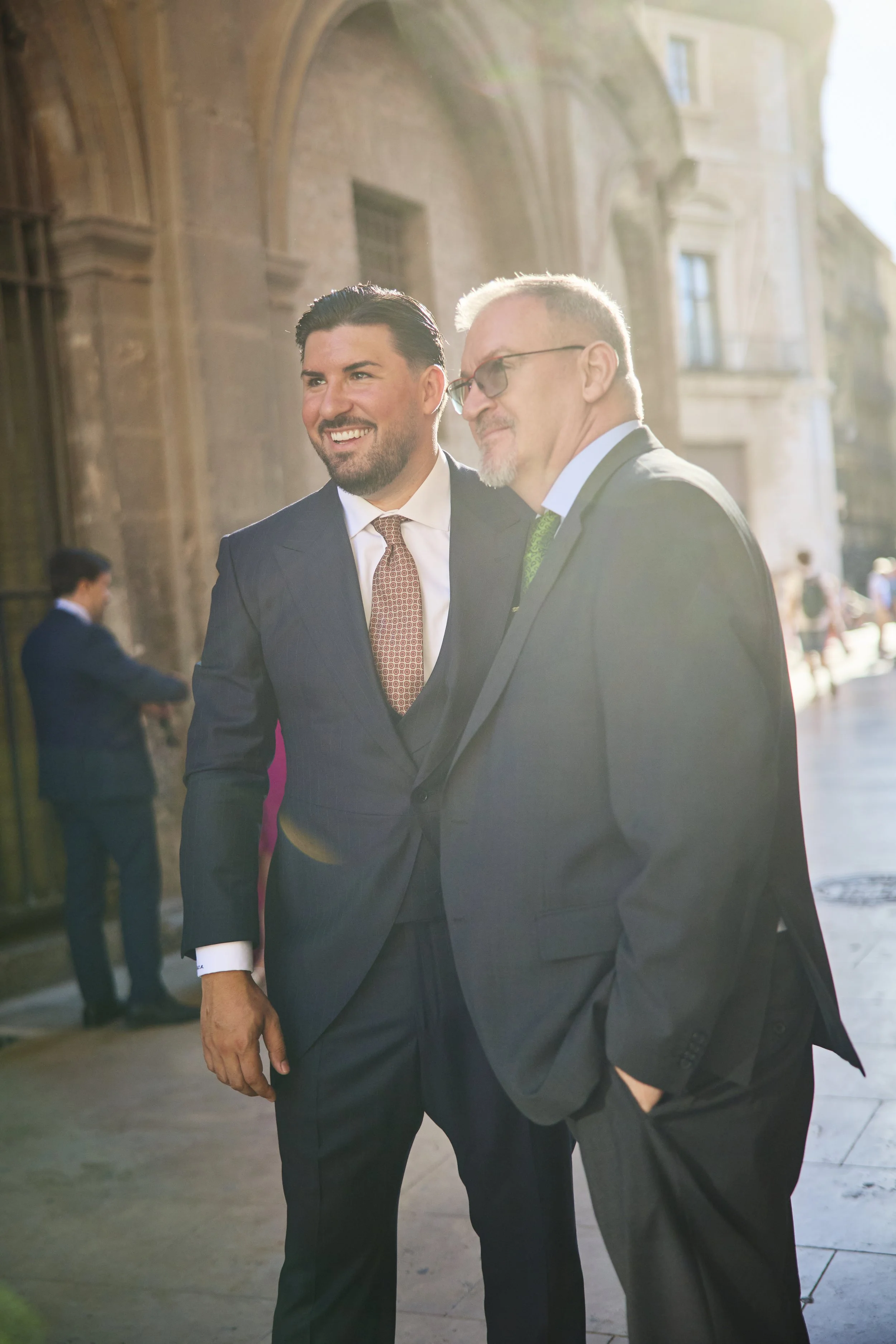 Dos hombres de negocios en trajes conversando en la calle, con edificio antiguo de piedra y ventanas detrás, al sol de la tarde.