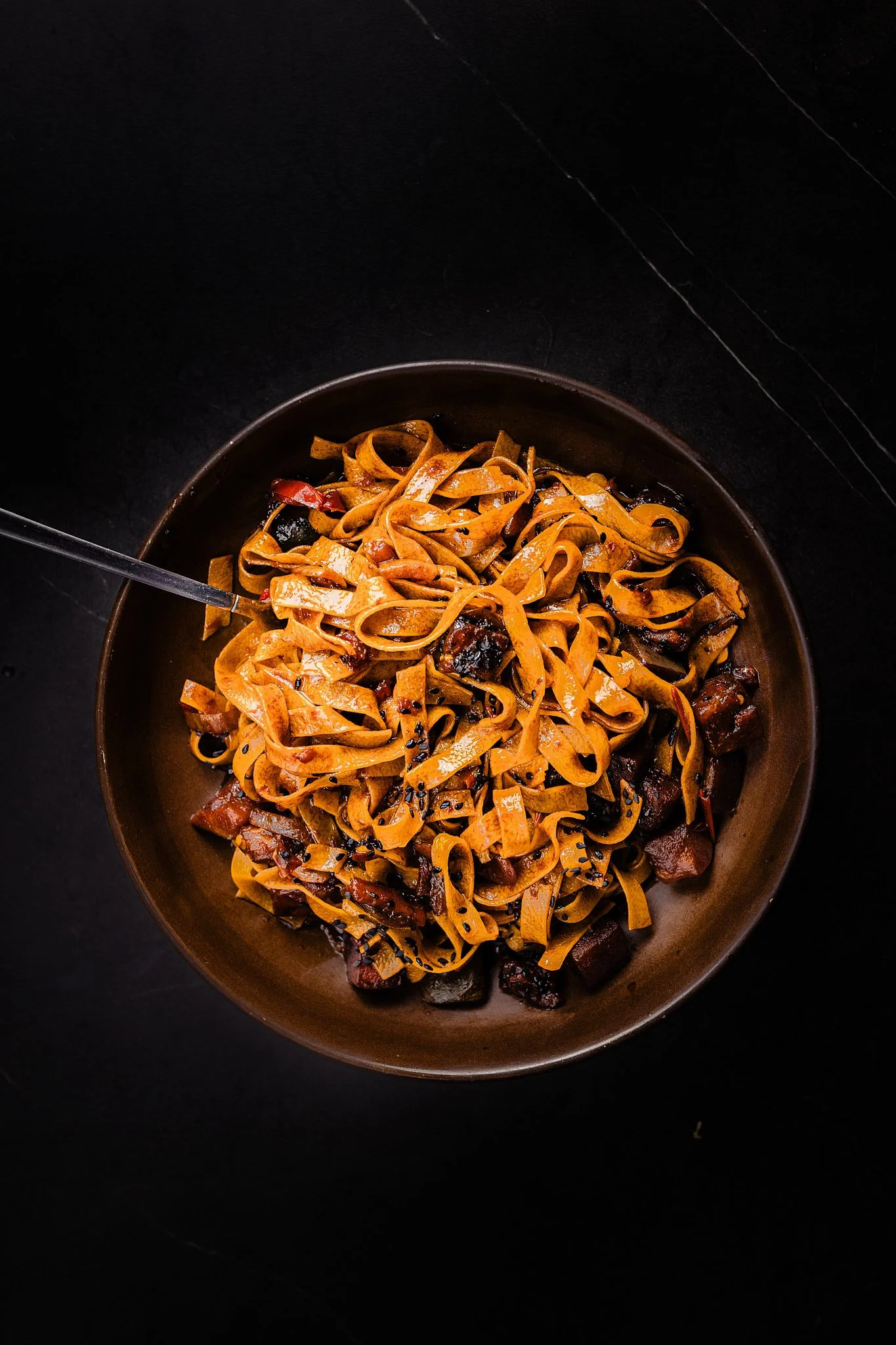 Plato de fideos frotados con sésamo negro y trozos de cerdo en un bol de color marrón oscuro, sobre una superficie negra.
