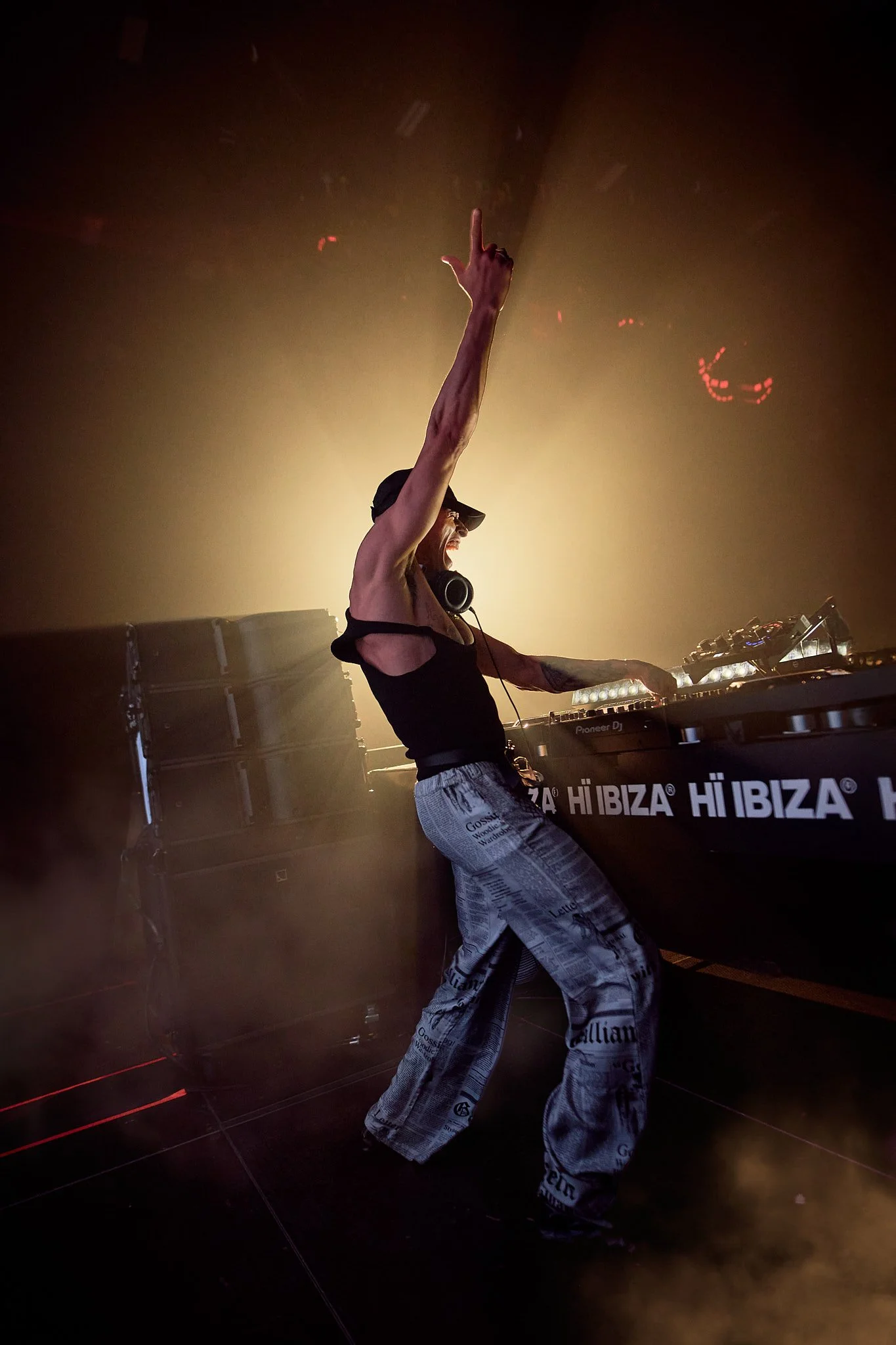 DJ en concierto con una mano levantada, rodeado de humo y luces, en un escenario oscuro.
