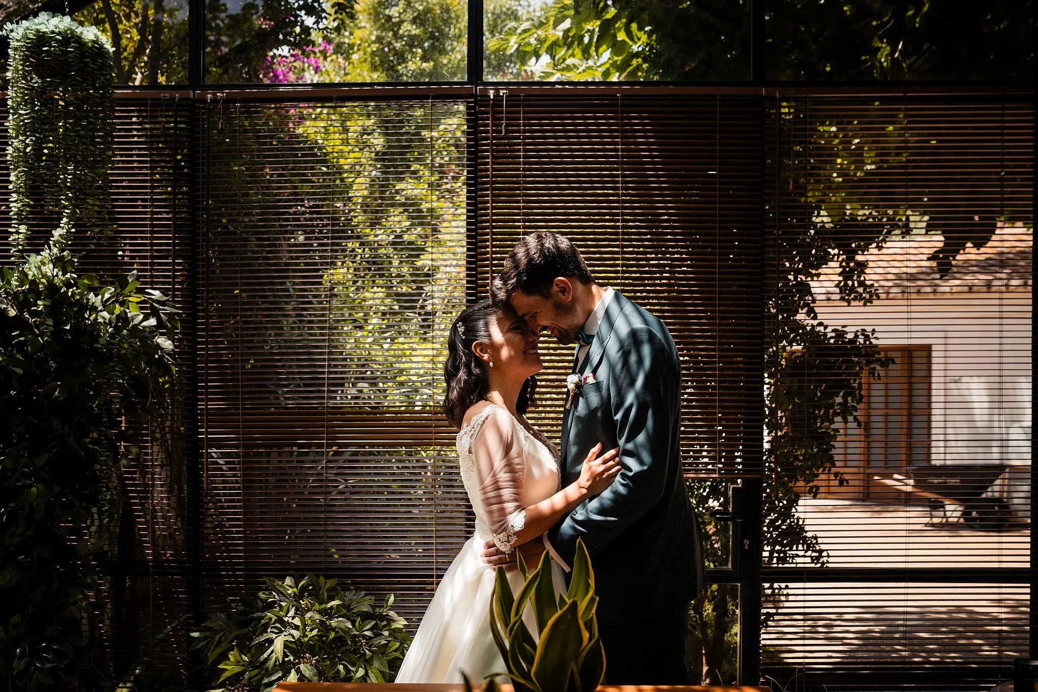 Pareja de recién casados en una boda, abrazados y sonriendo en un espacio interior con ventanas y plantas alrededor.