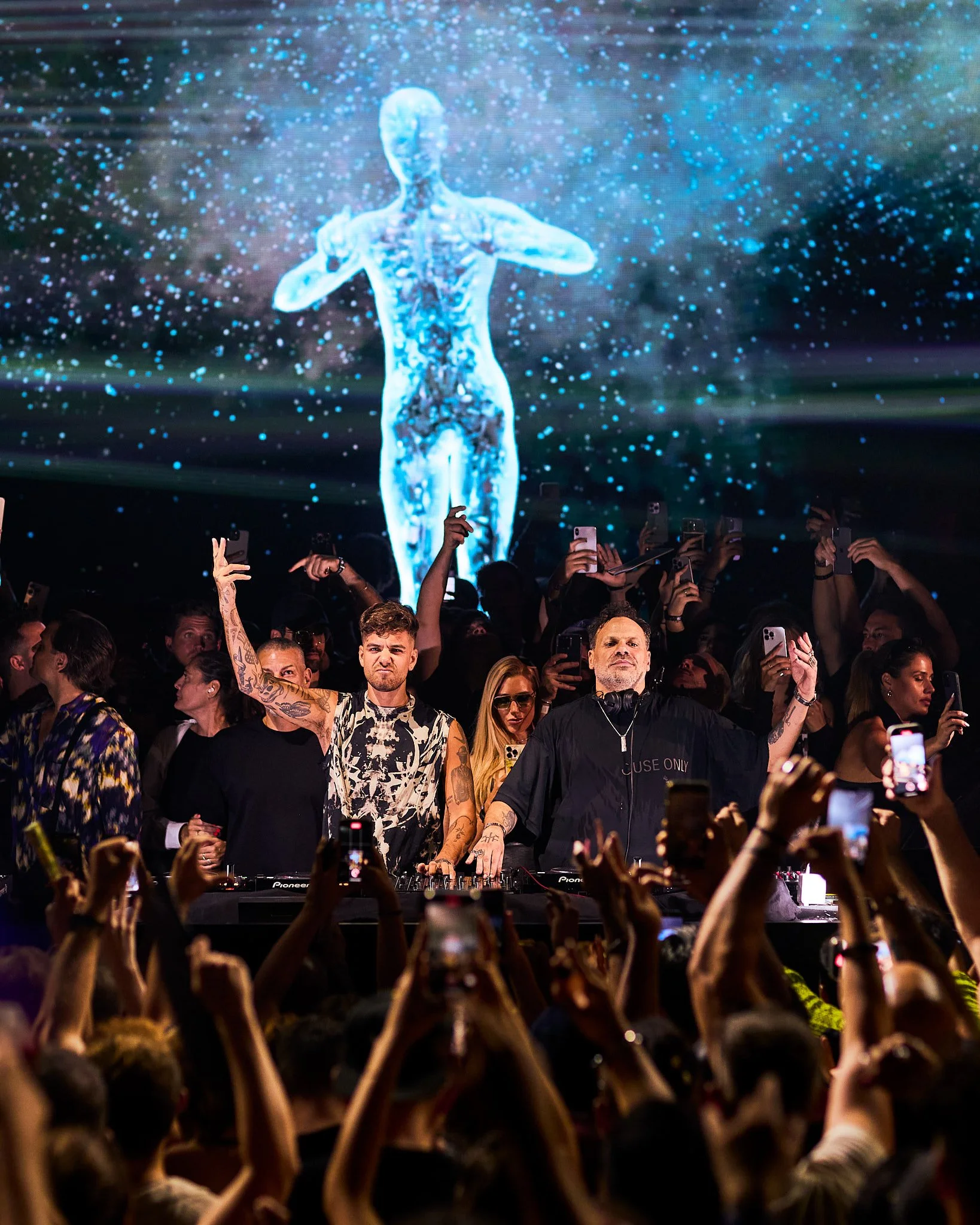 Afterlife, anyma y MRAK Un DJ y dos personas en un escenario con una pantalla que muestra una figura humana digital y un fondo de luces y efectos visuales, en medio de una multitud en un concierto.