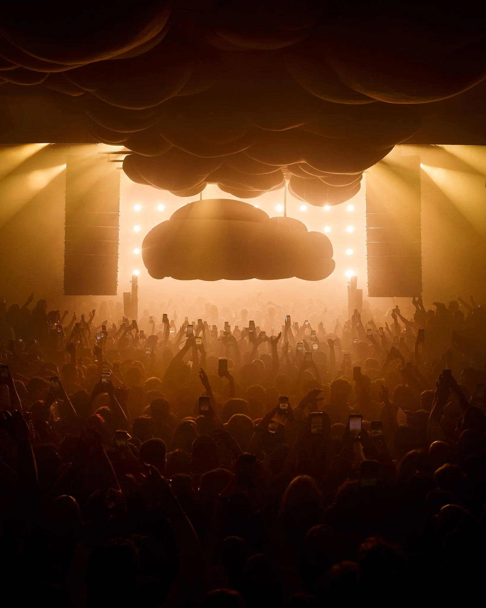 Concierto con muchas personas, escenario con siluetas, luces brillantes y nubes inflables decorativa en el techo.