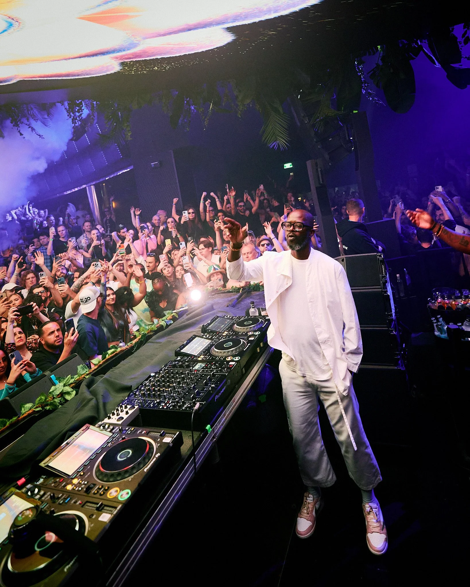 Un DJ tocando en un escenario con una multitud bailando y disfrutando en un club nocturno con iluminación colorida.