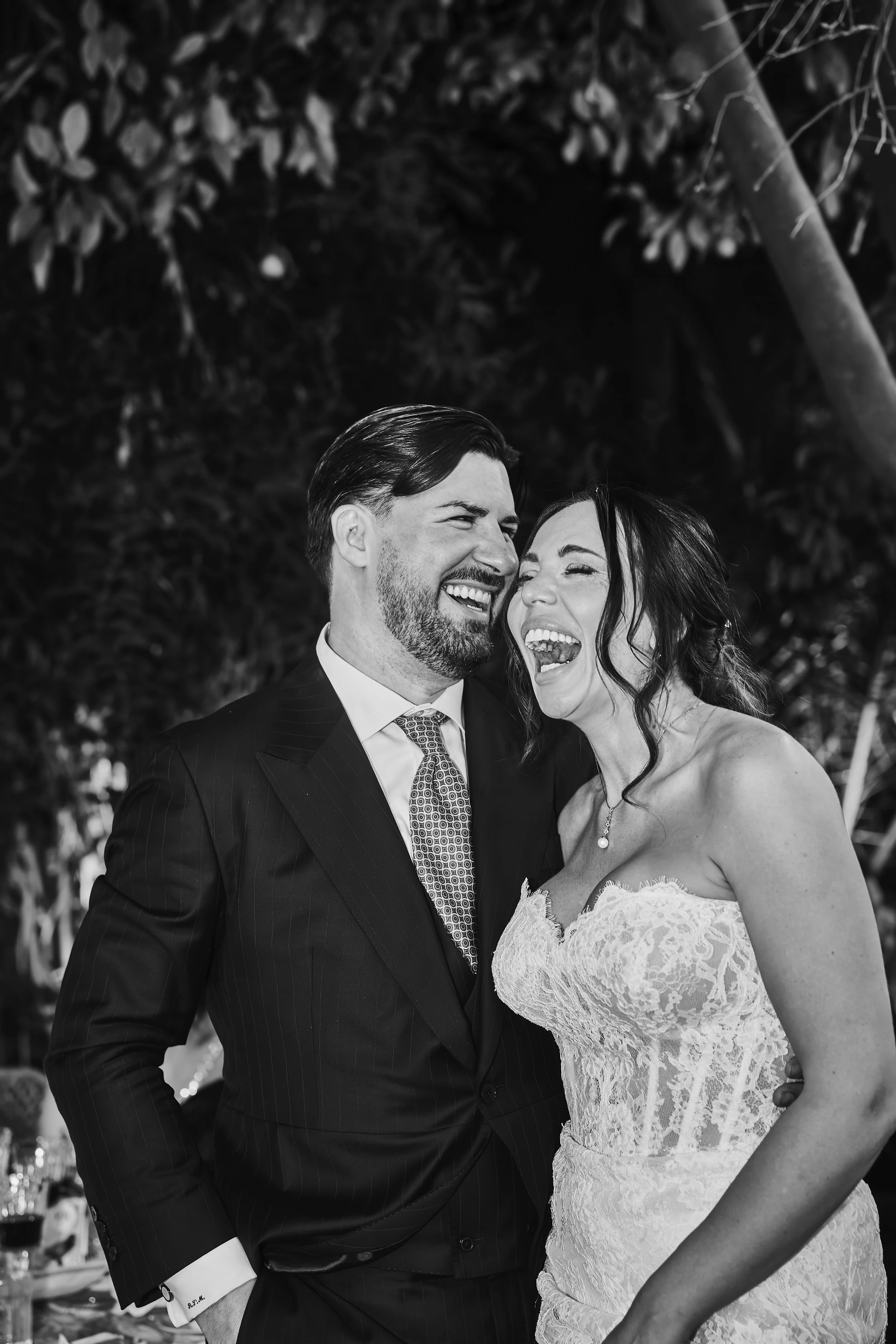 Pareja de boda riendo y abrazándose, el hombre viste traje oscuro y la mujer vestido de novia de encaje.