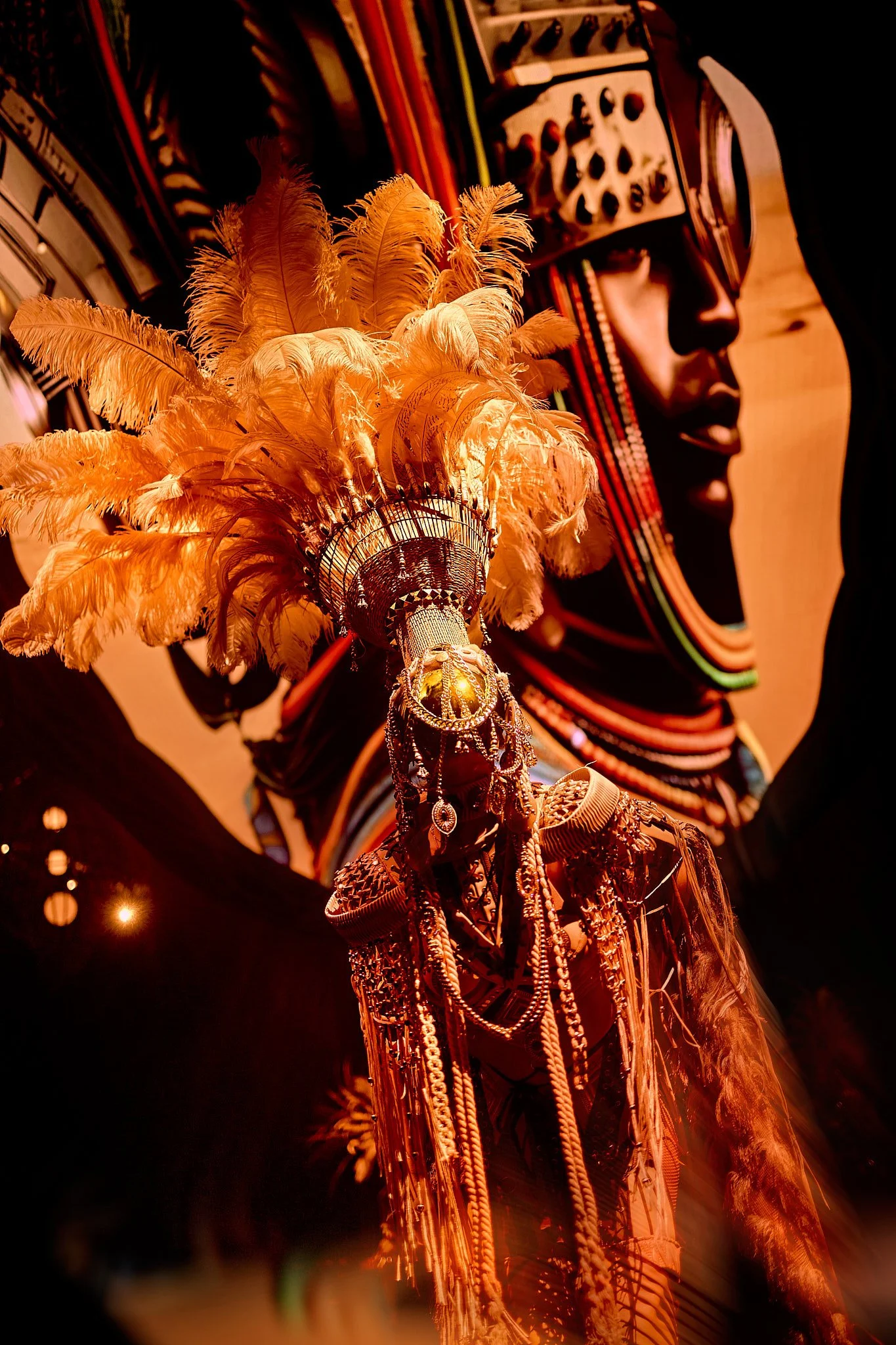 Una figura con joyas y plumas de colores cálidos, con un fondo de retrato artístico de una mujer con toques africanos en tonos oscuros.