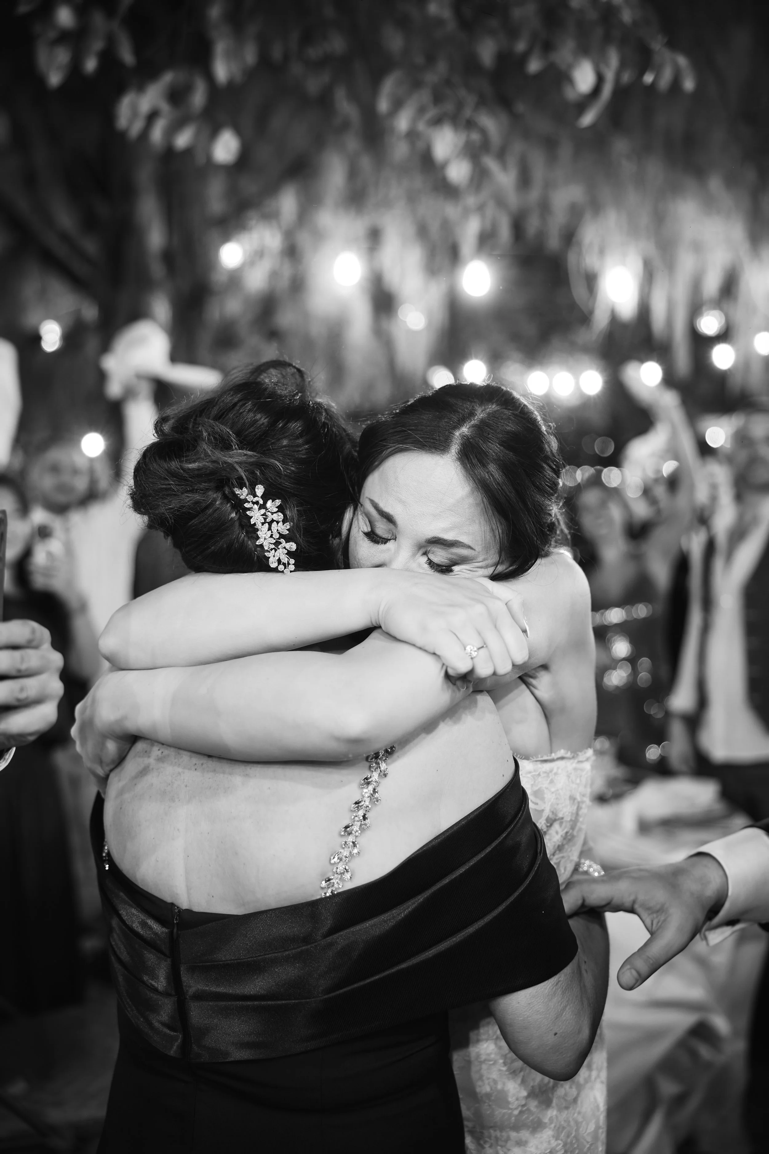 Dos mujeres abrazándose con lágrimas en los ojos en una celebración o evento especial, en un entorno con luces y otras personas en el fondo.