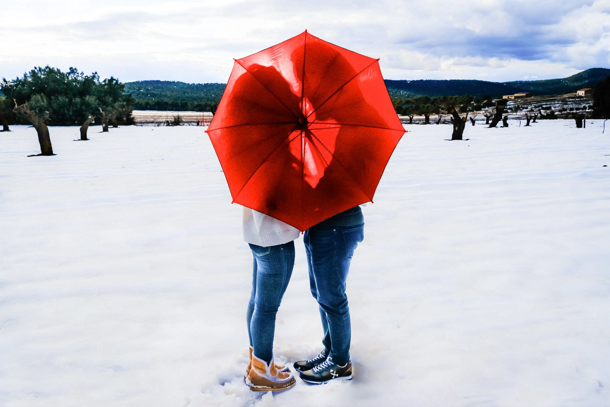 Una pareja abrazándose en un campo nevado con árboles podados y un cielo nublado, sosteniendo un paraguas rojo.