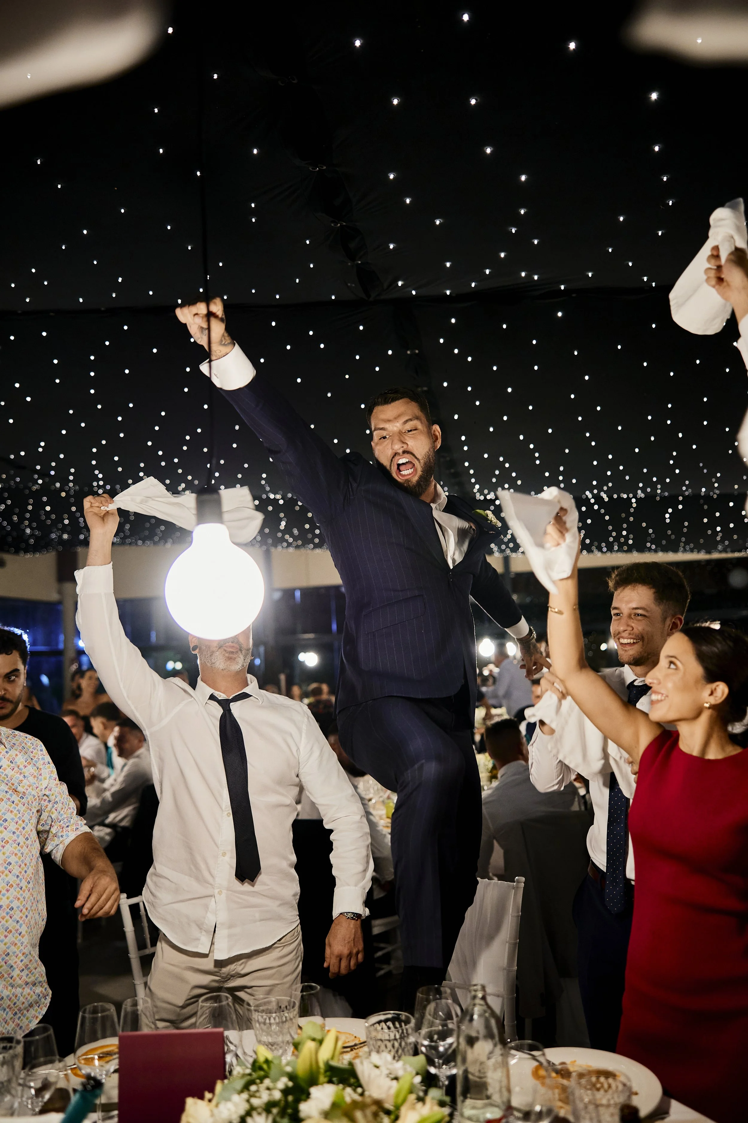 Hombres y mujeres en una celebración de noche, algunos sosteniendo servilletas en el aire, con un hombre vestido con traje de novio en el centro levantando un puño y mostrando ánimo.