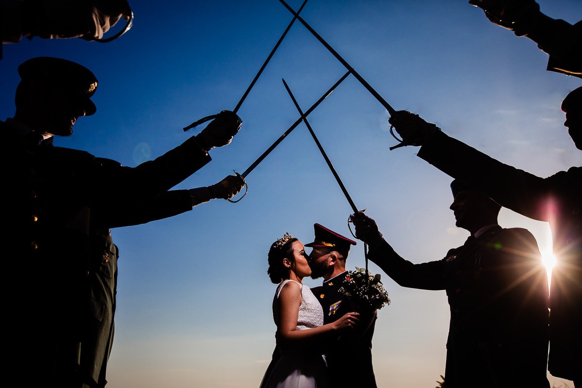 Silhouette de una pareja casada besándose, rodeada por soldados que levantan sus sables en forma de arco contra el cielo al atardecer.