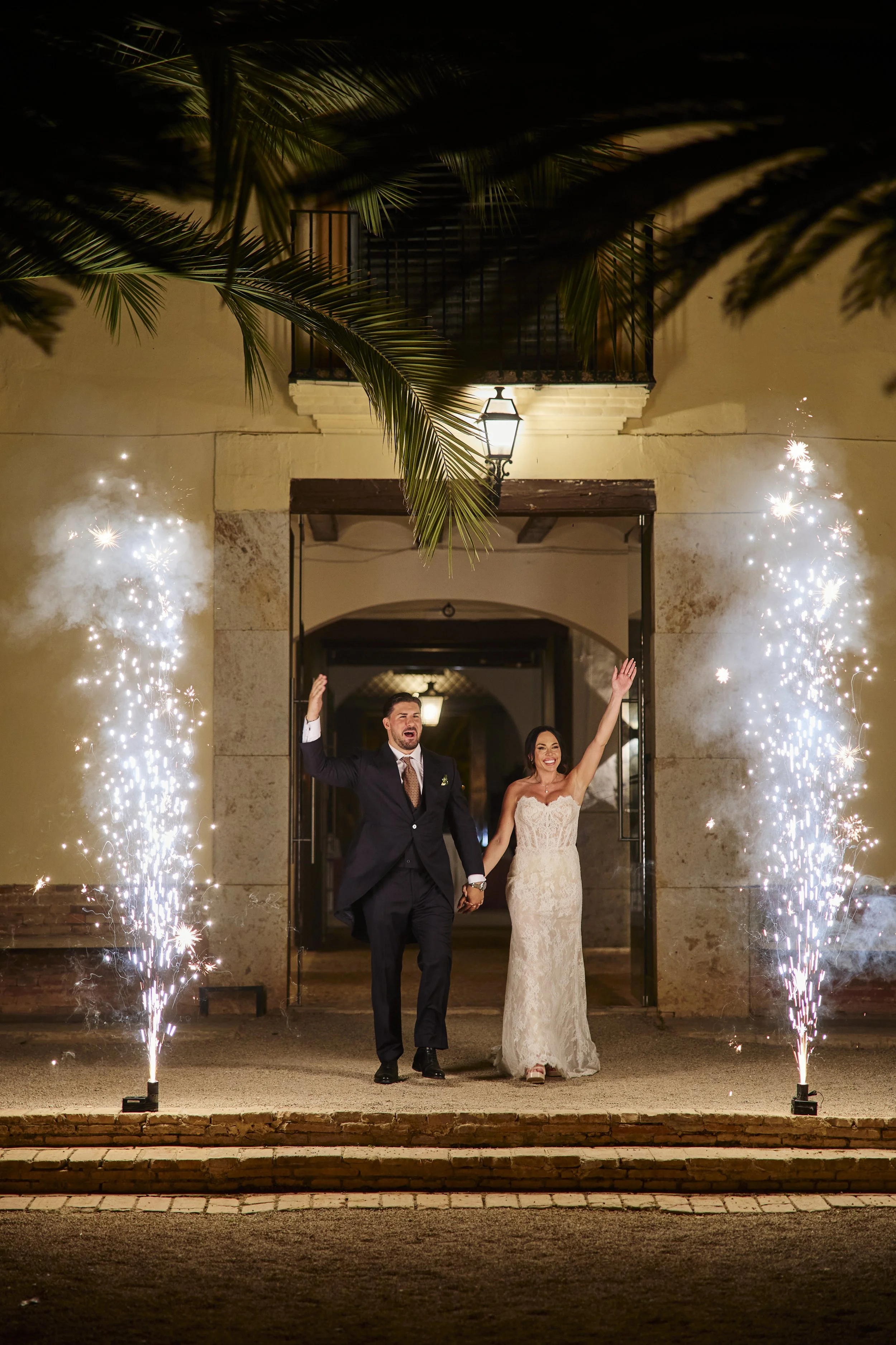 Pareja de recién casados caminando entre fuegos artificiales en la noche, celebrando su boda en la entrada de un edificio con farol y balcón
