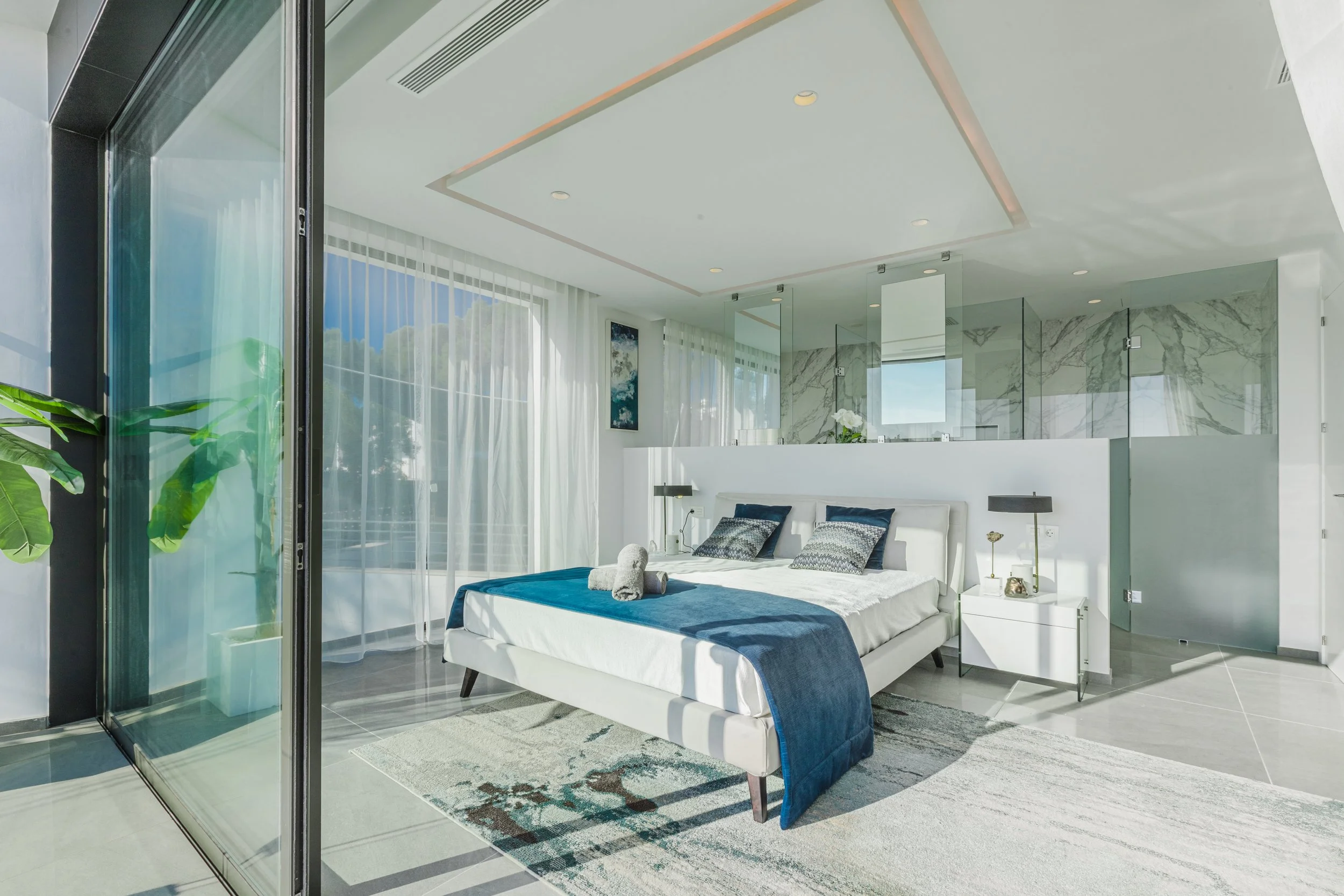 Habitación moderna con cama blanca, ropa de cama azul, cojines grises, y cortinas blancas, con ventanas grandes y detalles en mármol y vidrio.