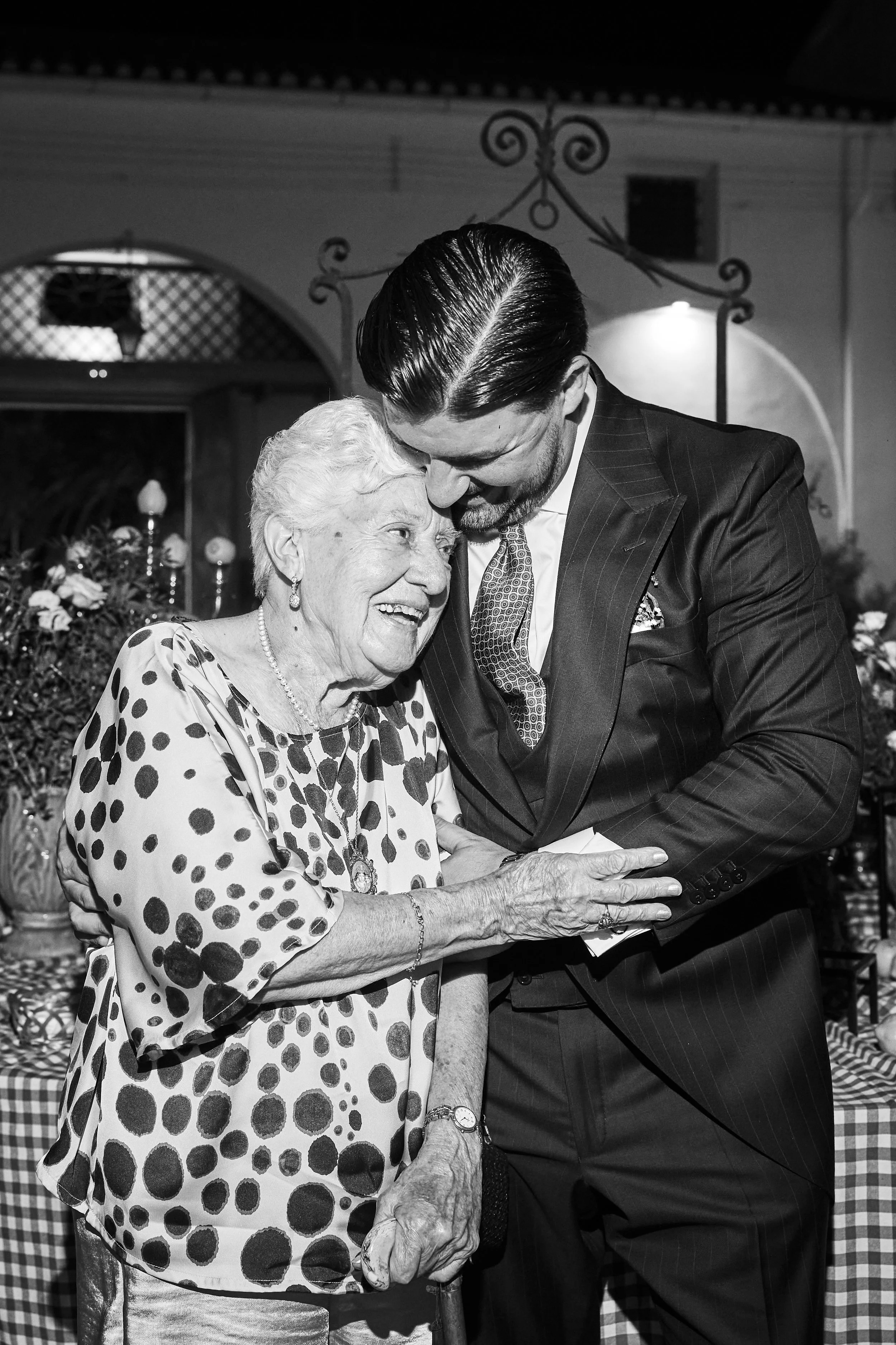 Un hombre joven con cabellera brillante y traje oscuro abrazando y acariciando a una anciana con blusa de lunares. Están sonriendo y parecen felices en un ambiente decorado con flores y velas.