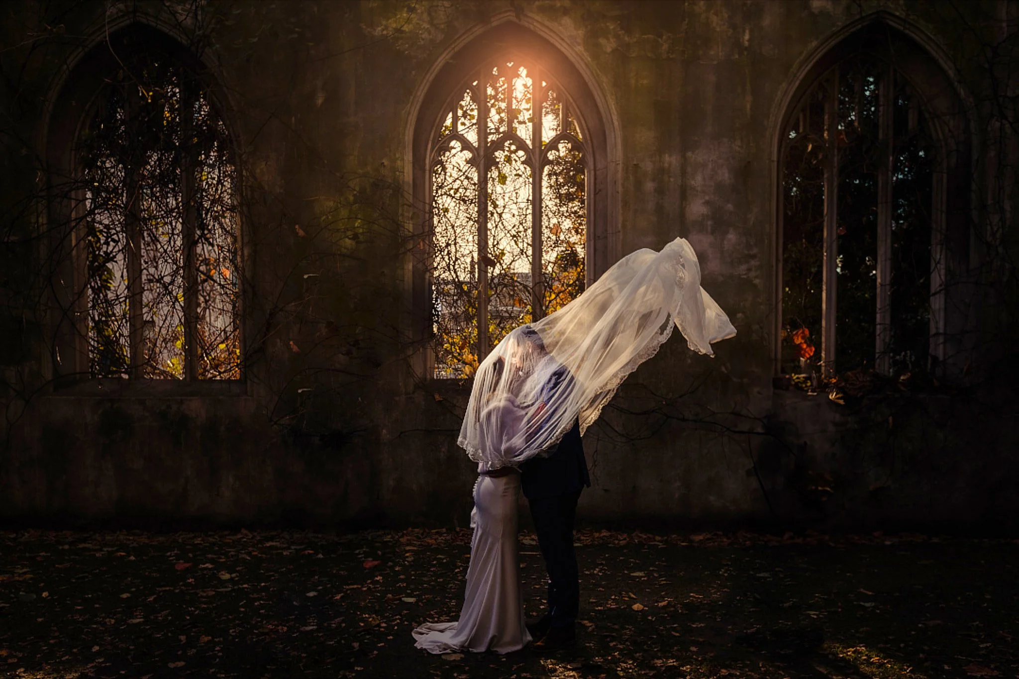 Pareja de novios en un lugar abandonado y oscuro, con ventanas y paredes cubiertas de vides y hojas secas, en una fotografía con luz cálida y romántica que crea una atmósfera misteriosa y melancólica.