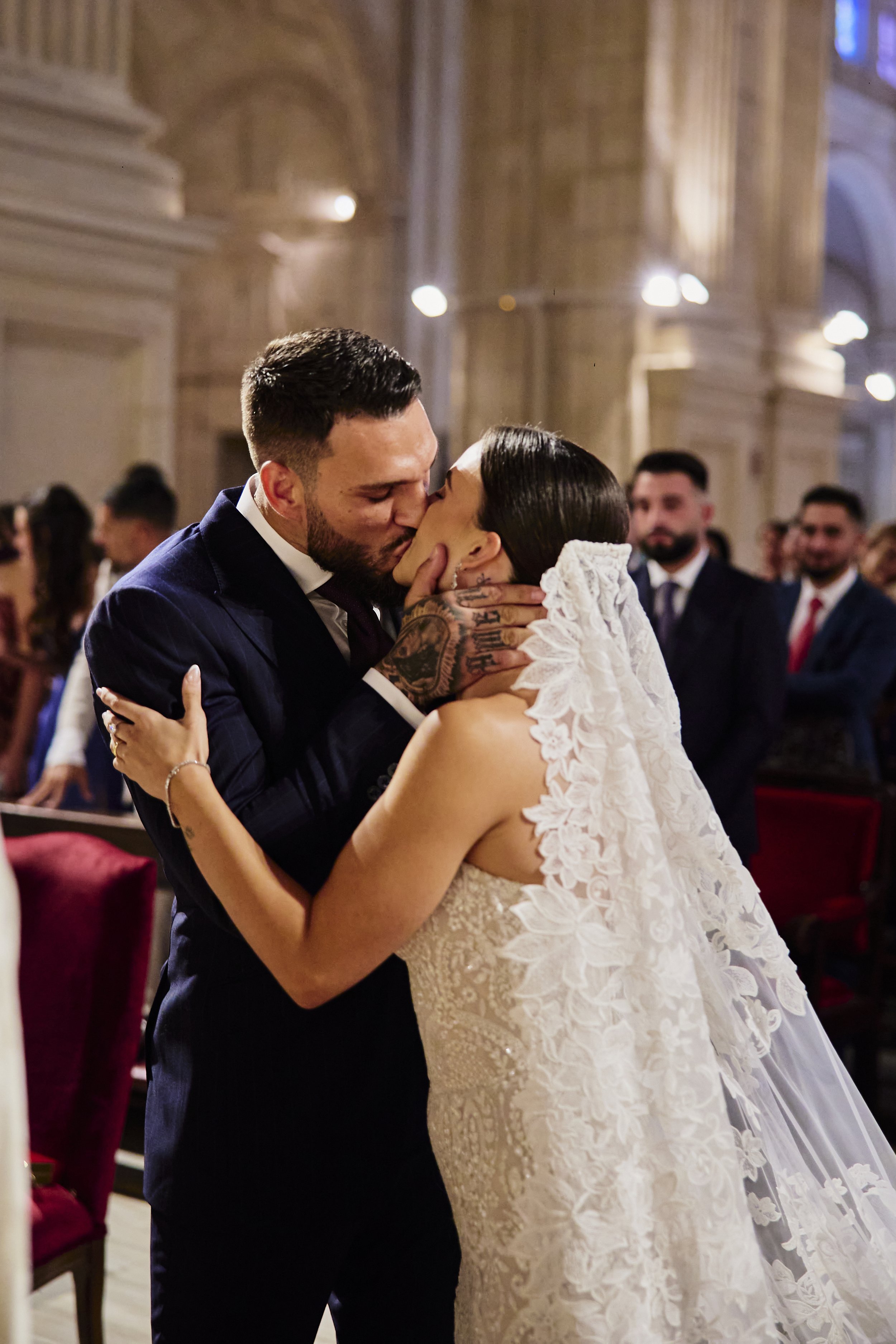 Una pareja en su boda compartiendo un beso en una iglesia, con invitados al fondo observando.