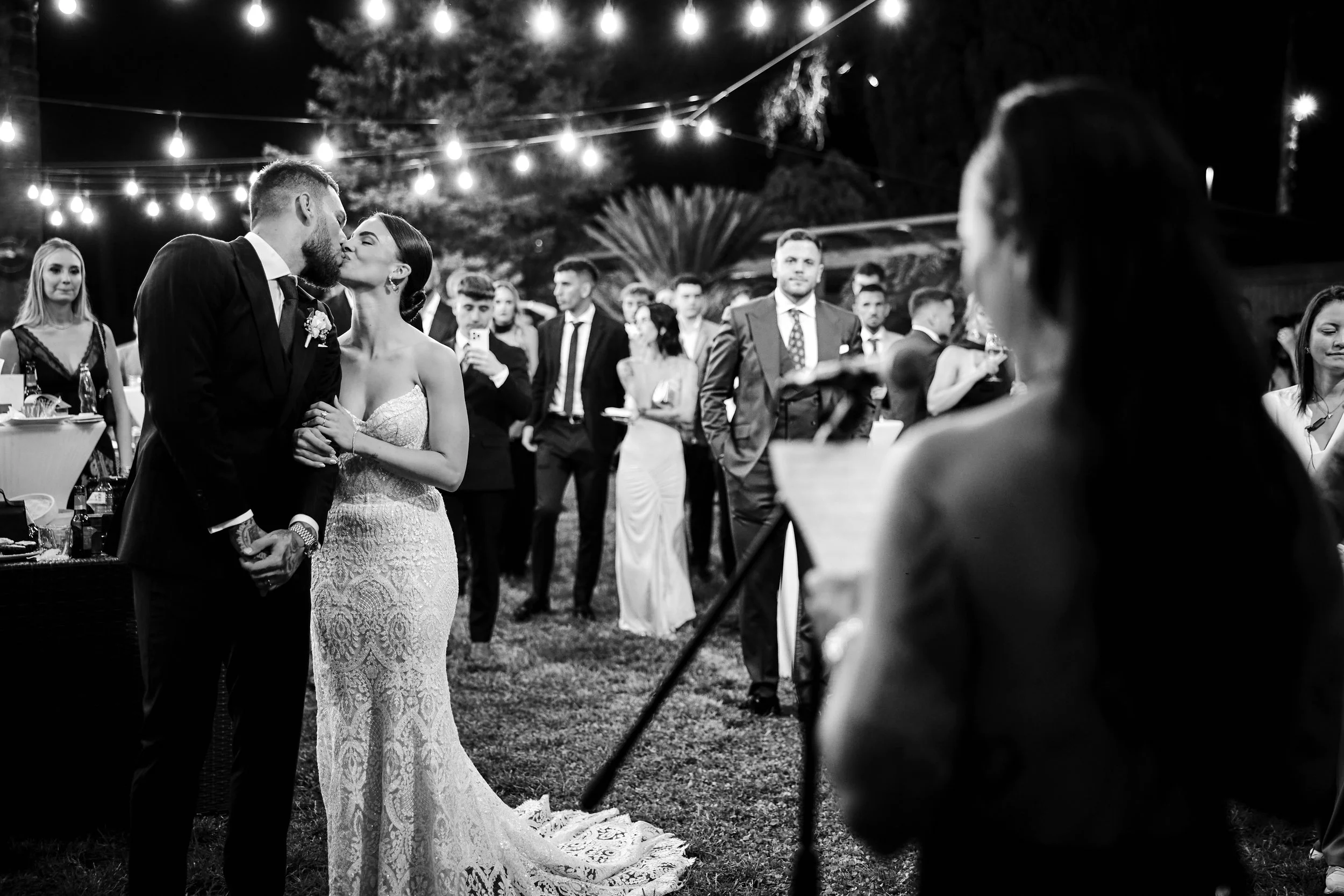 Pareja de recién casados besándose en una boda en exteriores, con invitados observando y tiras de luces decorativas en el fondo.