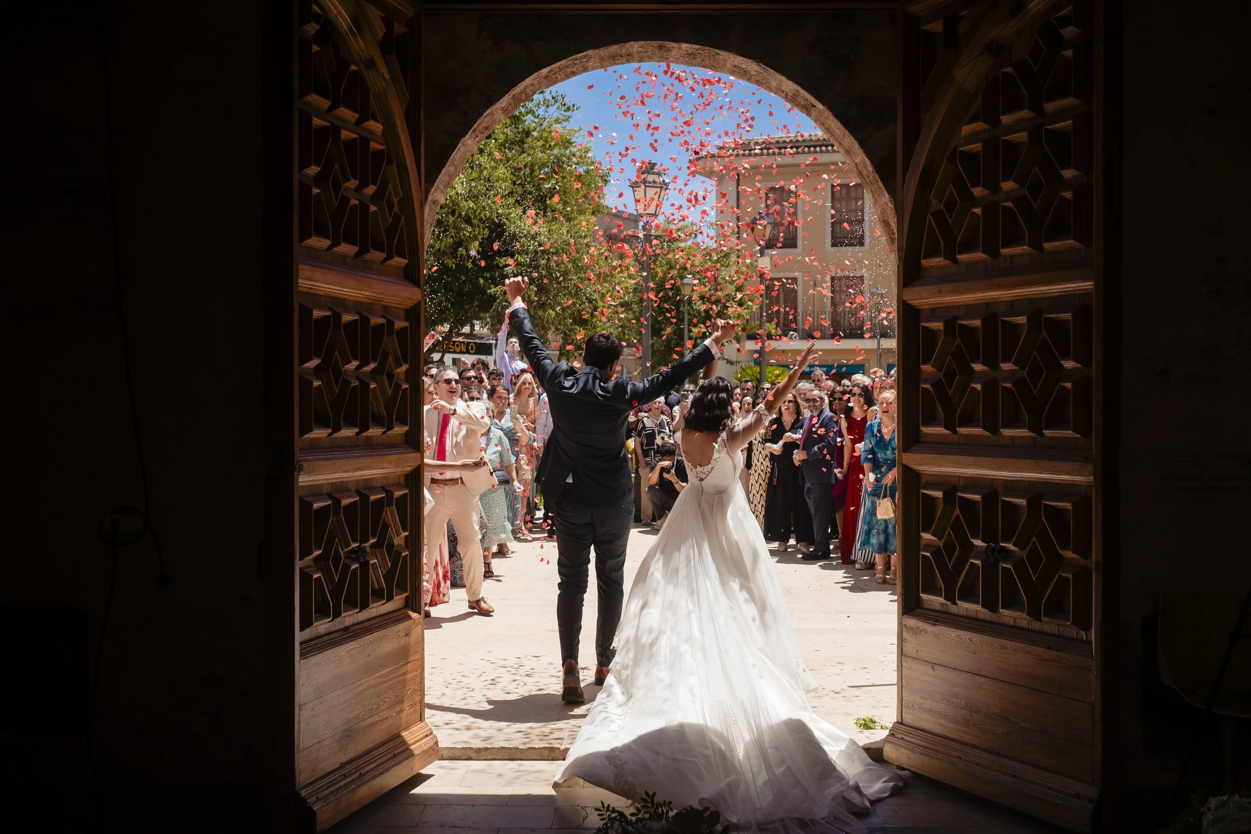 Pareja de novios celebrando su boda con invitados lanzando pétalos de rosa, vista desde la puerta de un edificio con ventanas de madera tallada, en un día soleado en una plaza urbana.
