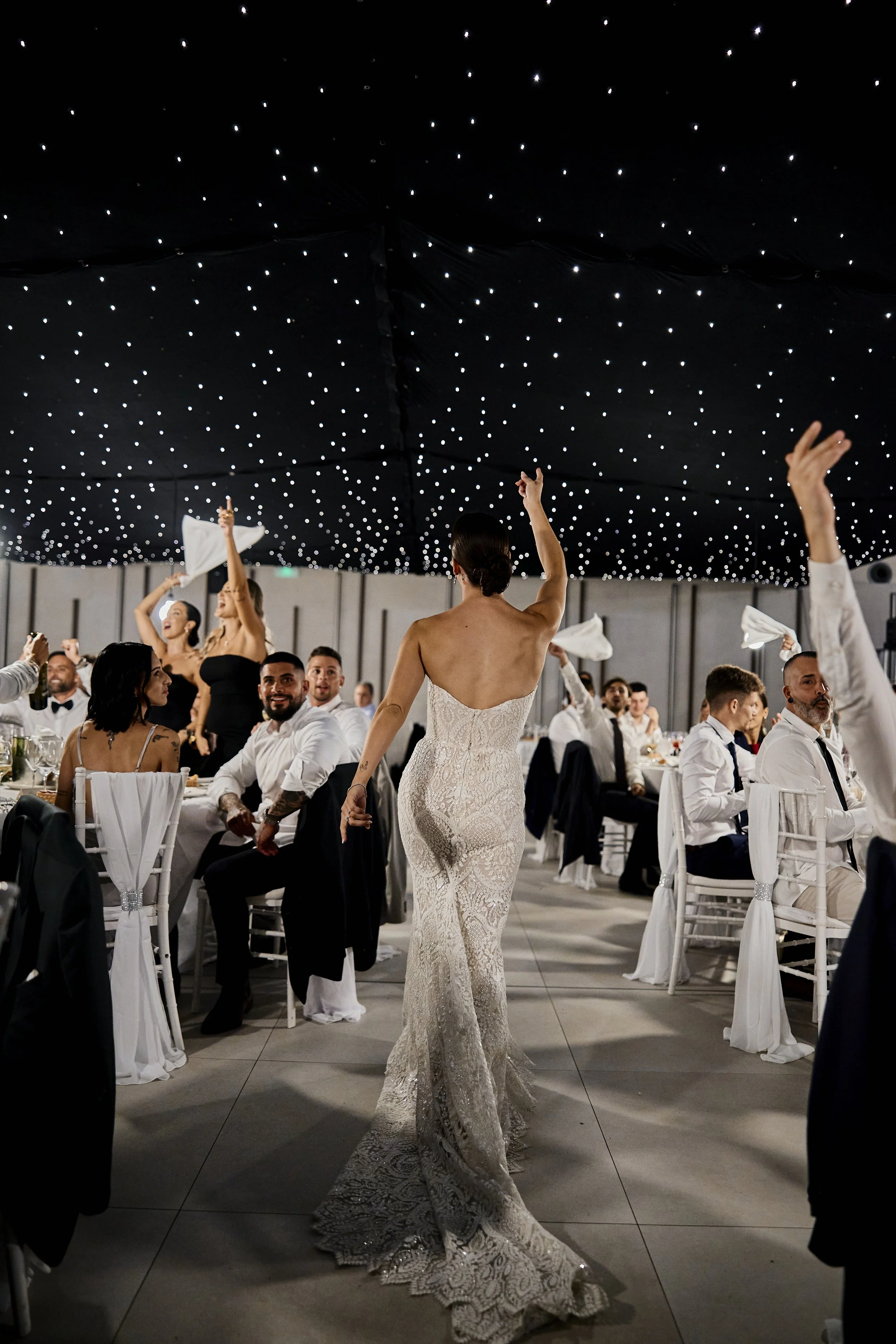 Mujer con vestido de novia en un baile en un evento elegante, rodeada de personas sentadas en mesas, con techo decorado con luces pequeñas.