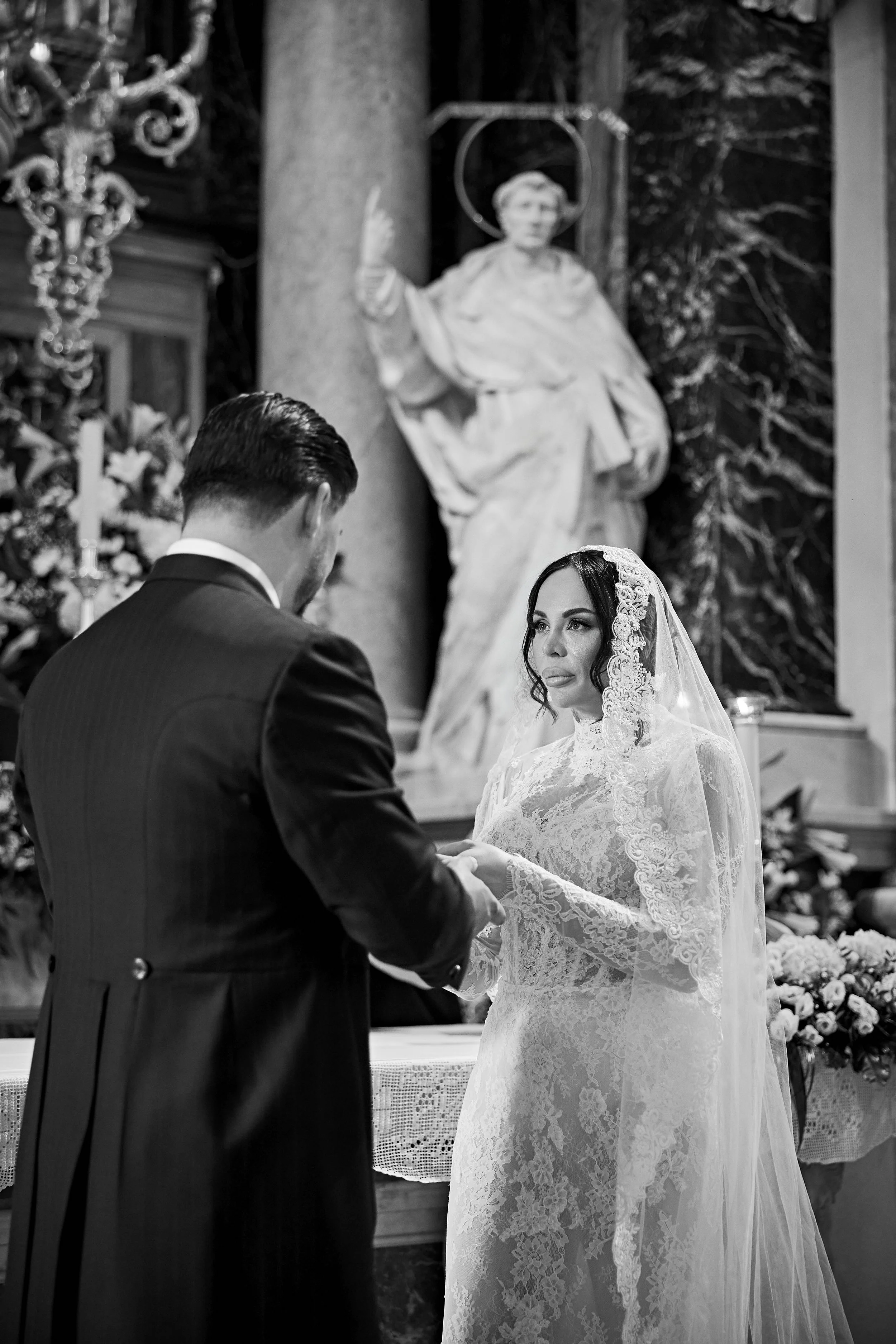 Una pareja en una boda religiosa, la mujer con vestido de novia y velo, el hombre con traje, en una iglesia con una estatua religiosa al fondo.