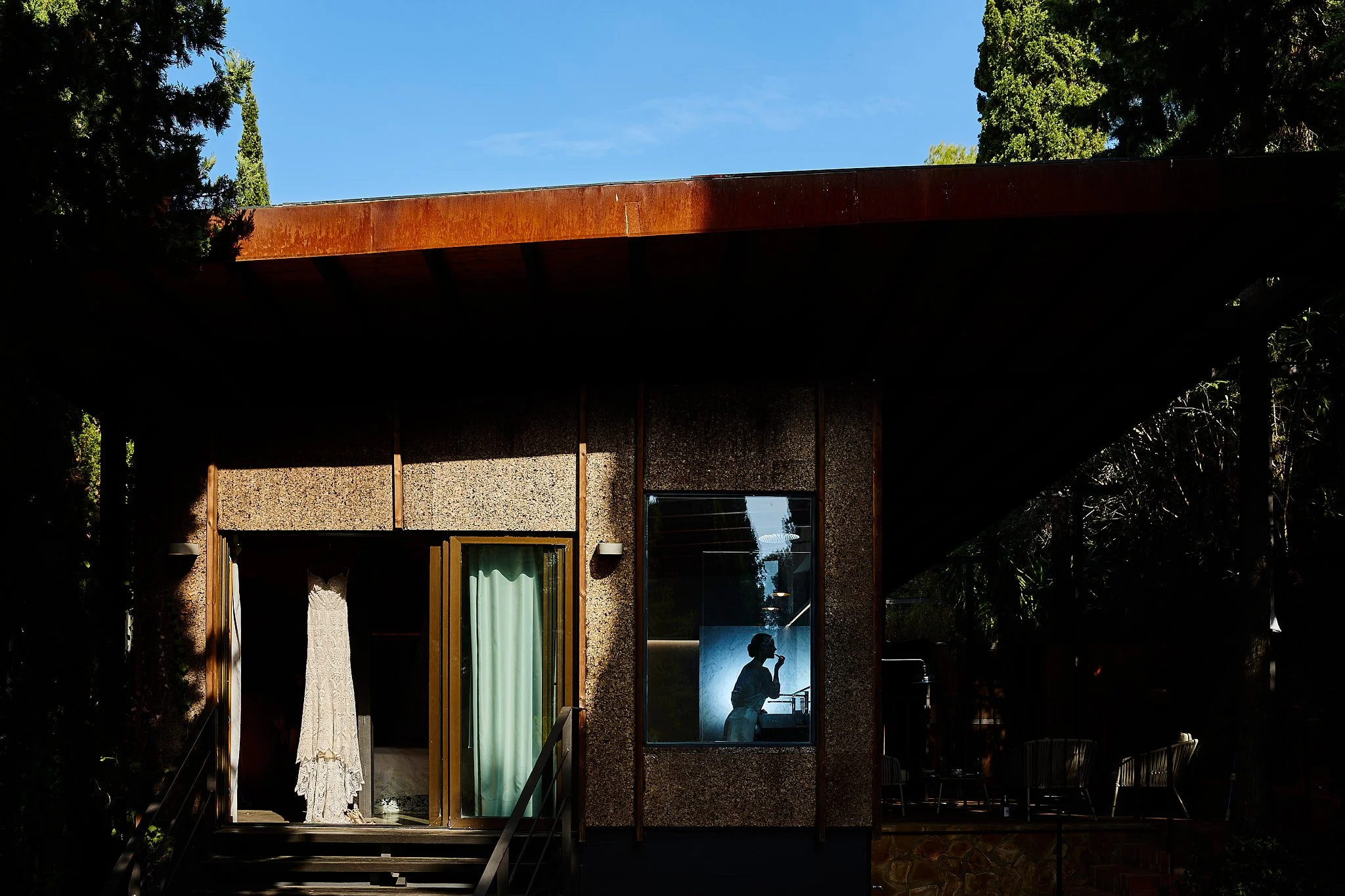 Una casa moderna de un piso con ventanas grandes, en la ventana del interior se ve la silueta de una mujer con el cabello recogido, haciendo una pausa y sosteniendo algo en la mano, con paredes de color azul y un escritorio.