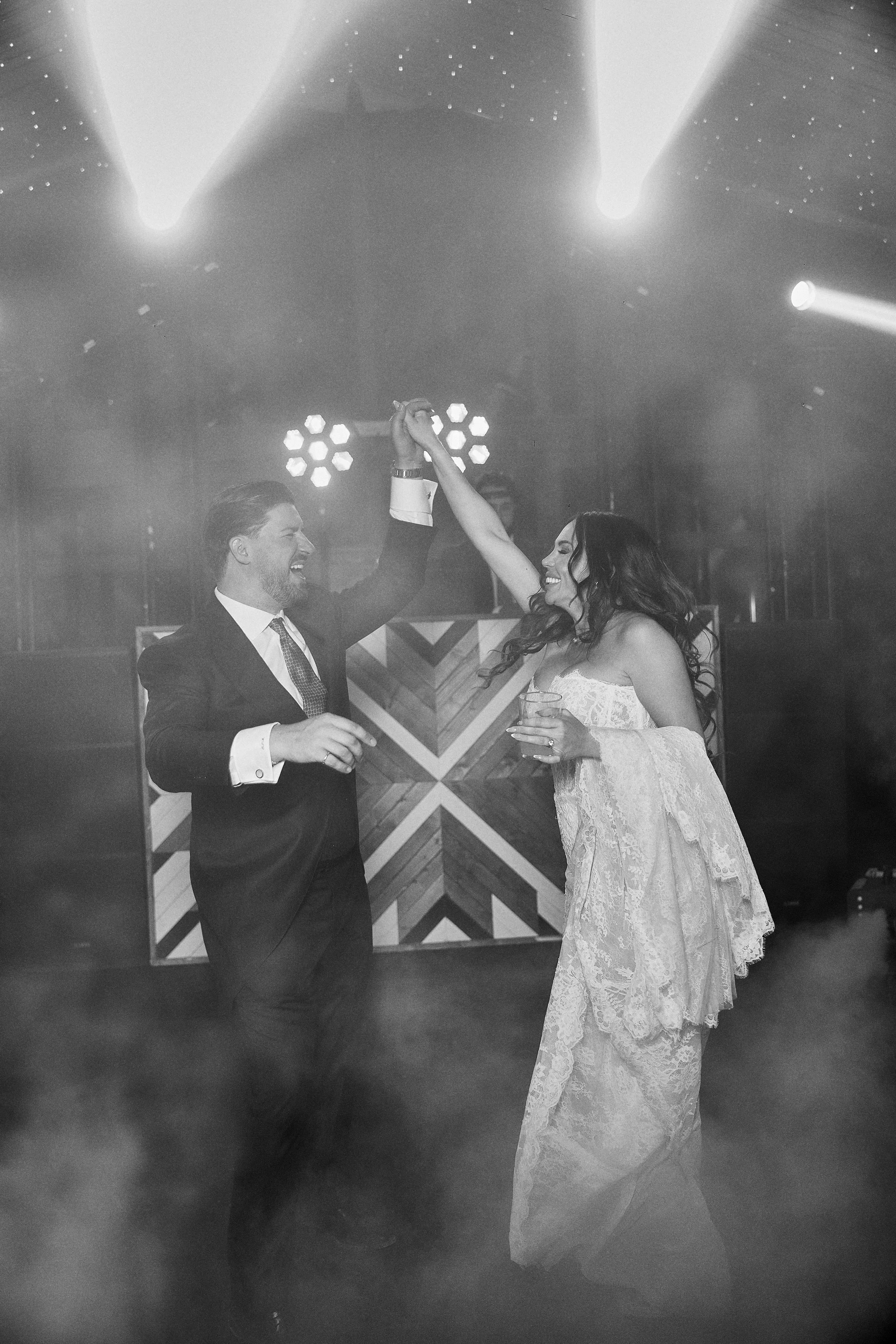 Pareja bailando en un evento, el hombre viste un traje negro y la mujer lleva un vestido largo de encaje y sostiene un vaso, ambos sonríen y se miran mientras bailan bajo luces brillantes.