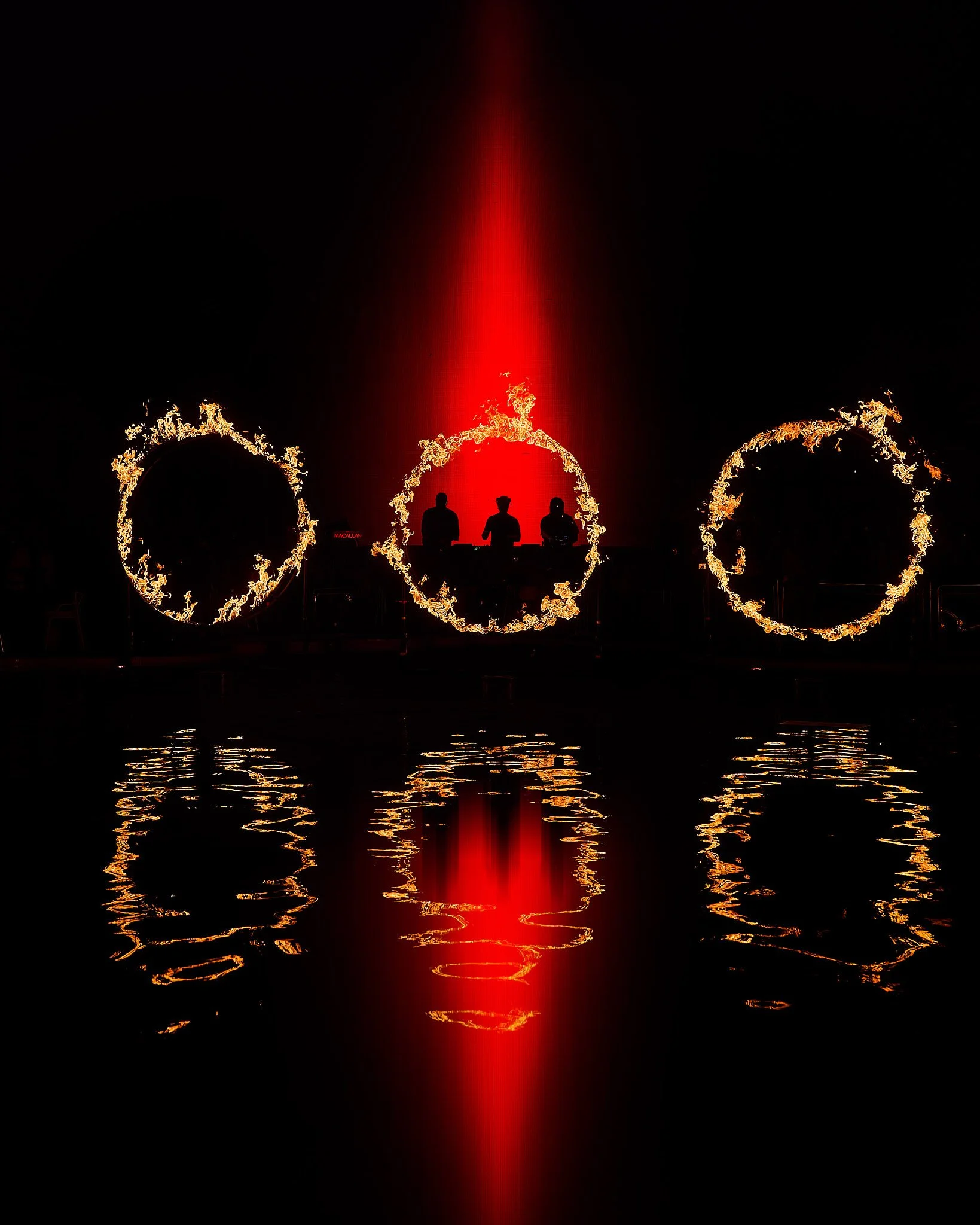 SWEDISH HOUSE MAFIA Escenario con tres círculos de fuego y tres figuras humanas en silueta, reflejándose en un río, con un fondo oscuro y una columna de luz roja.