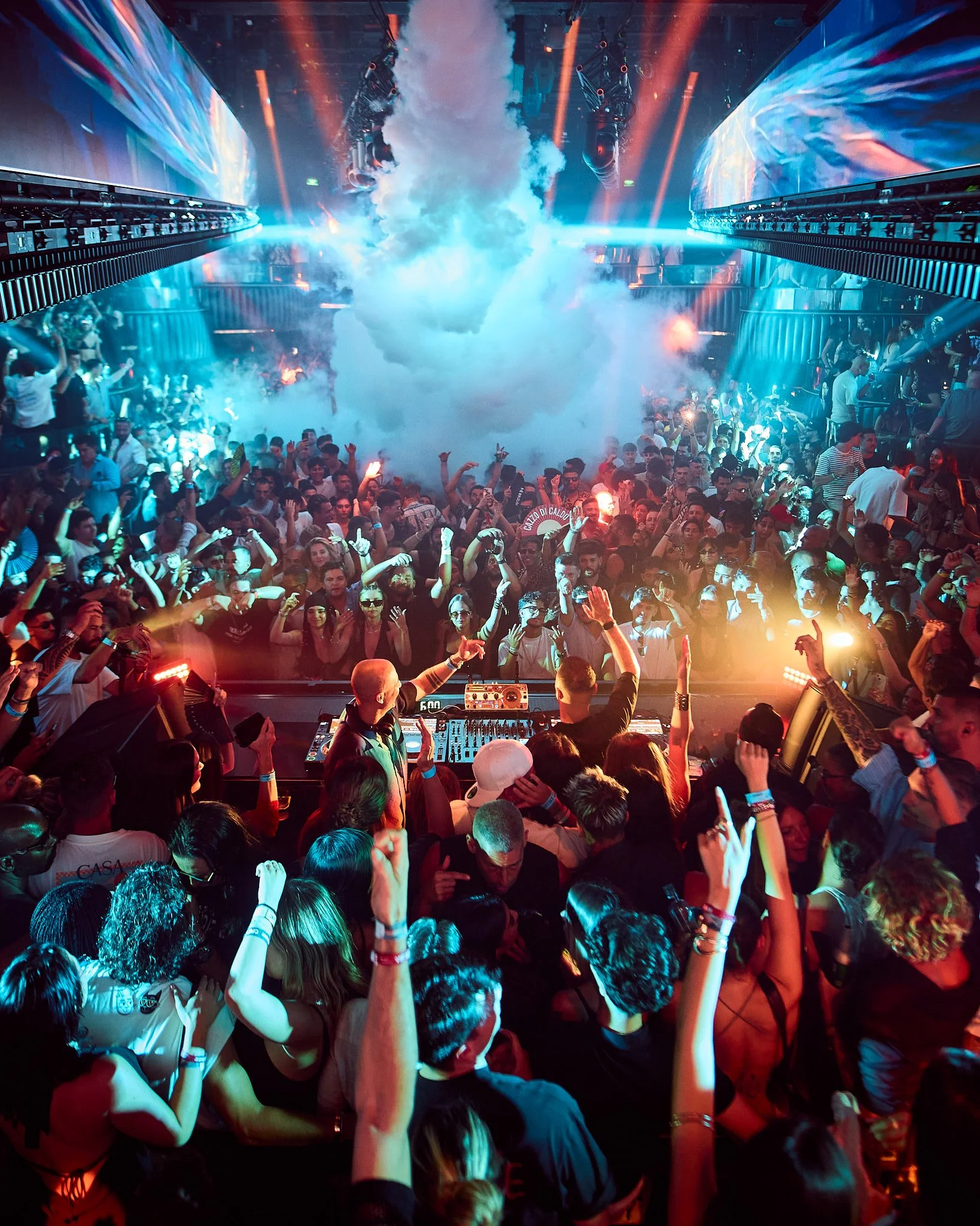 Un concierto en un club nocturno con una multitud bailando frente a un DJ en el escenario, con humo y luces de colores brillantes.