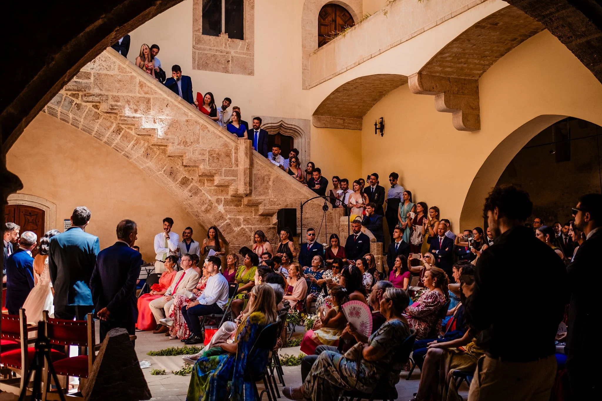 Una pareja de recién casados entrando a una iglesia para su boda, vista desde la entrada, con una multitud de invitados afuera y un edificio antiguo en el fondo; en blanco y negro.