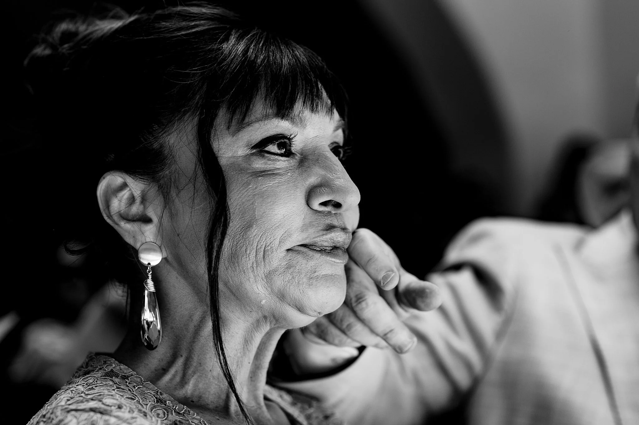 Retrato en blanco y negro de una mujer mayor con pendientes largos, mirando hacia arriba y a la izquierda mientras alguien la acaricia en la mejilla.