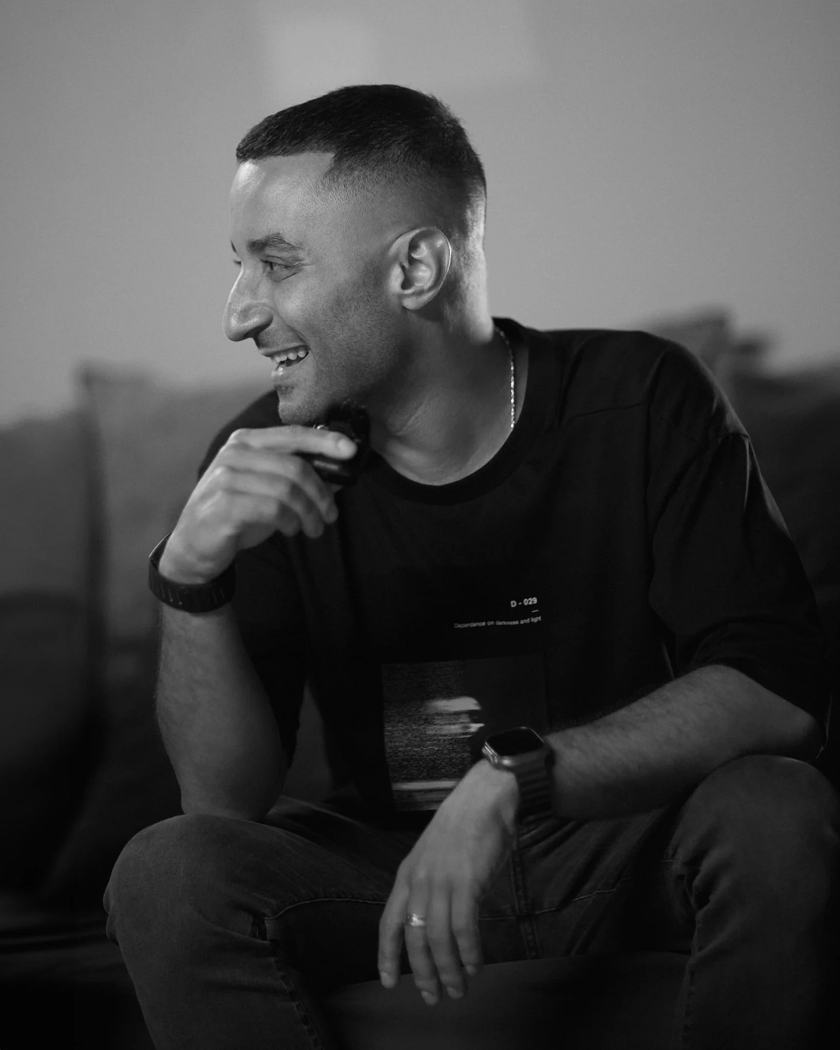 Un hombre sonriendo, sentado en un sofá, usando una camiseta negra y un reloj inteligente en la muñeca izquierda.